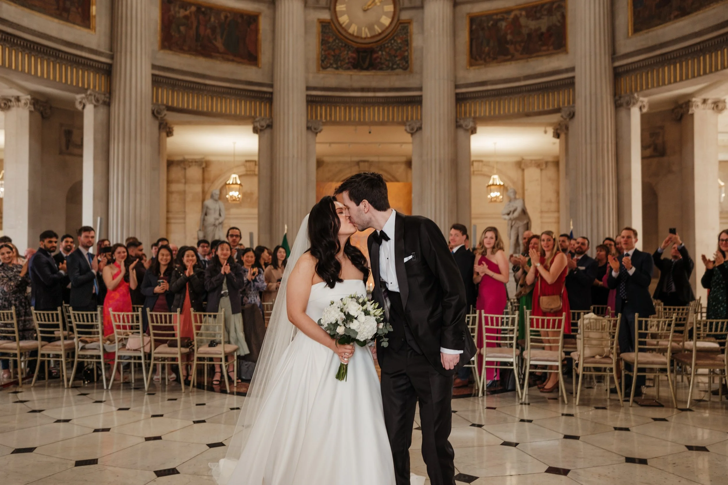 A bride and groom sharing a kiss during their wedding ceremony in a grand hall with tall columns and statue backgrounds, surrounded by seated and standing guests.