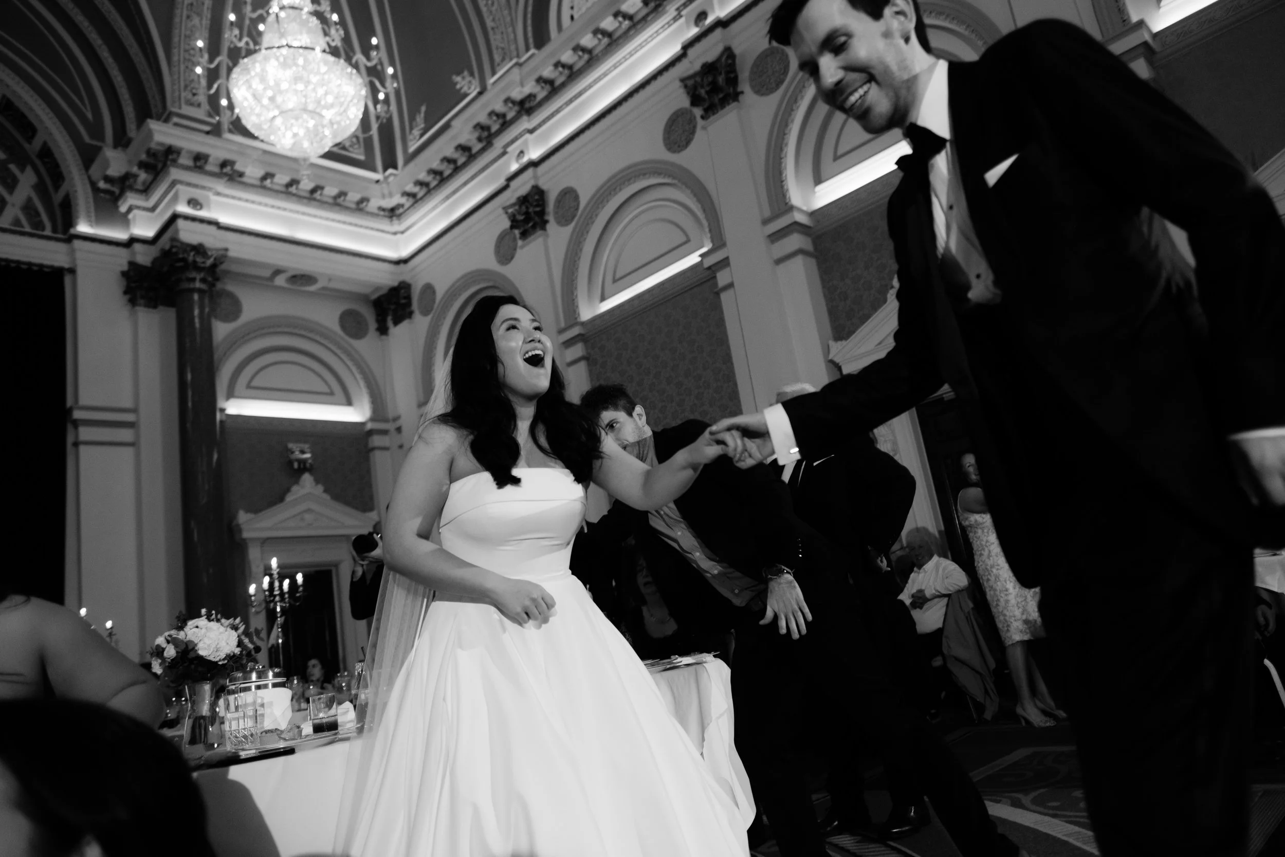 A joyful bride and groom dancing and laughing during their wedding reception in an elegant, ornate hall with chandeliers.