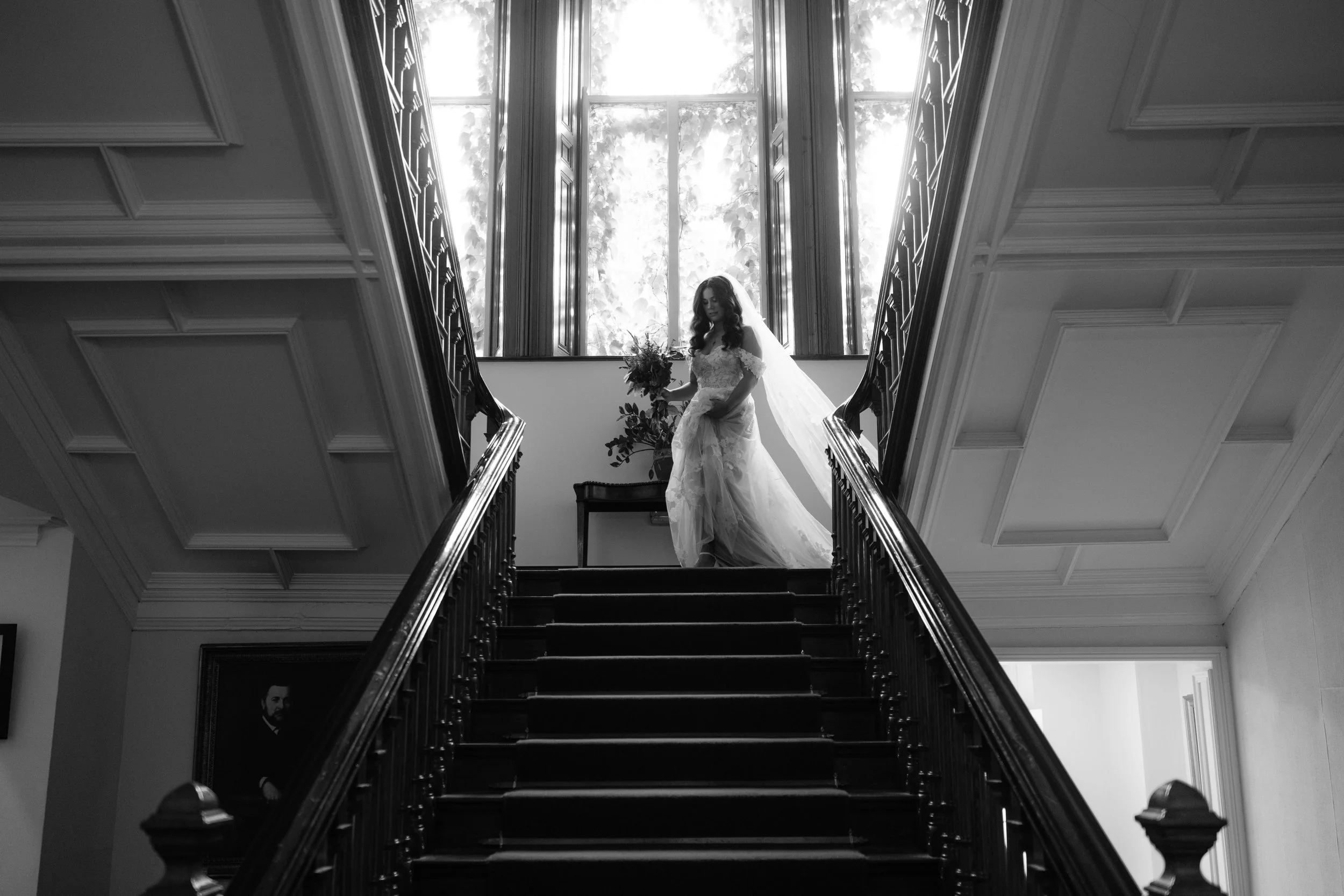 A bride in a wedding dress holding a bouquet, standing at the top of a staircase inside a house, with sunlight coming through large windows behind her.
