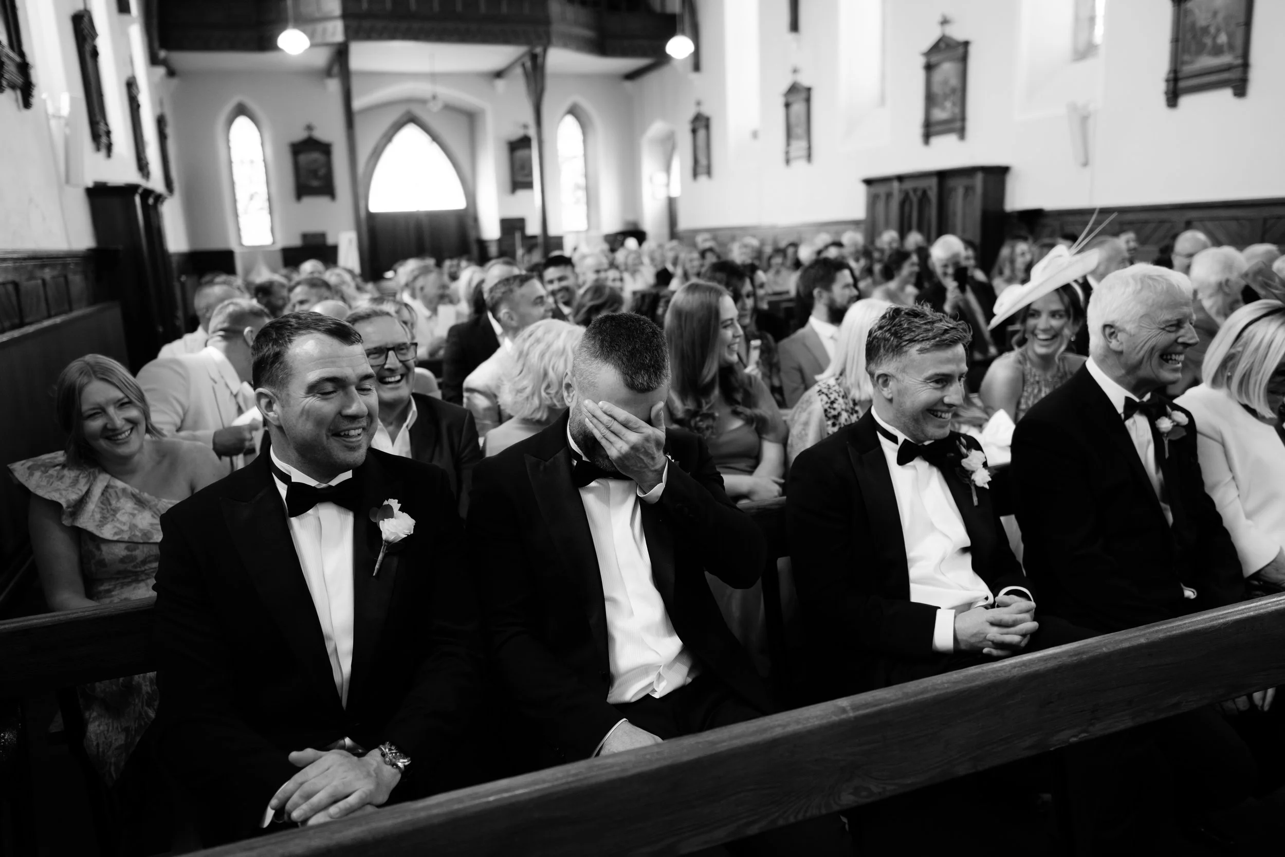A group of people attending a wedding ceremony inside a church, dressed in formal attire, with some men in tuxedos and women in elegant dresses, sitting and laughing.