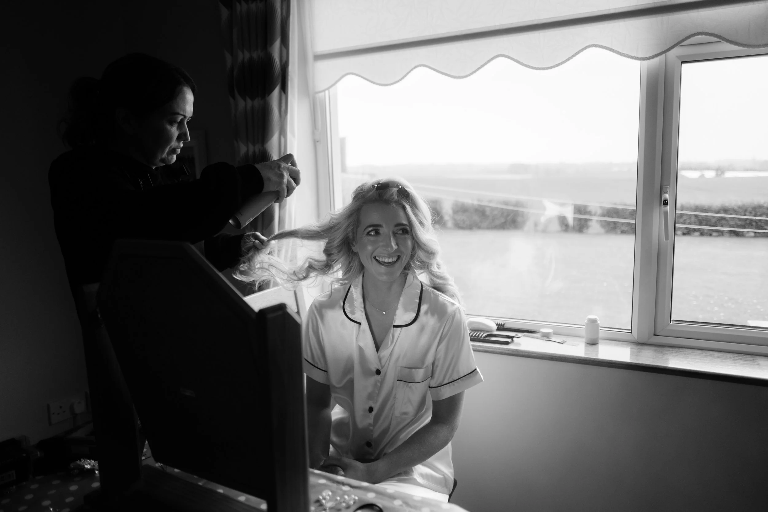 A woman sitting in front of a window with a violin smiling, while a hairstylist styles her hair during a photoshoot.