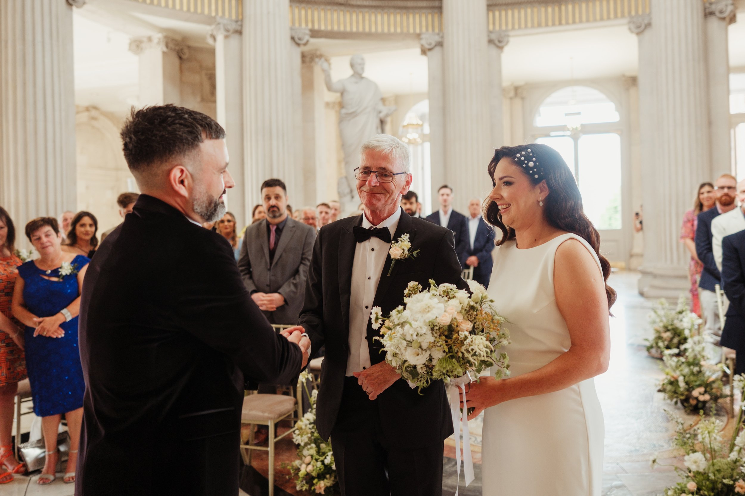 A wedding ceremony in a grand hall with tall columns, featuring a bride in a white dress holding a bouquet, a groom in a black suit, and an officiant in a tuxedo, surrounded by seated guests.