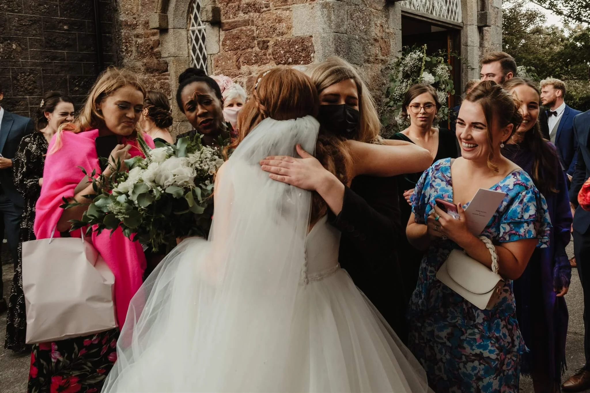 Group of women hugging and smiling at a wedding celebration outside a brick building, with some holding flowers and others taking photos.