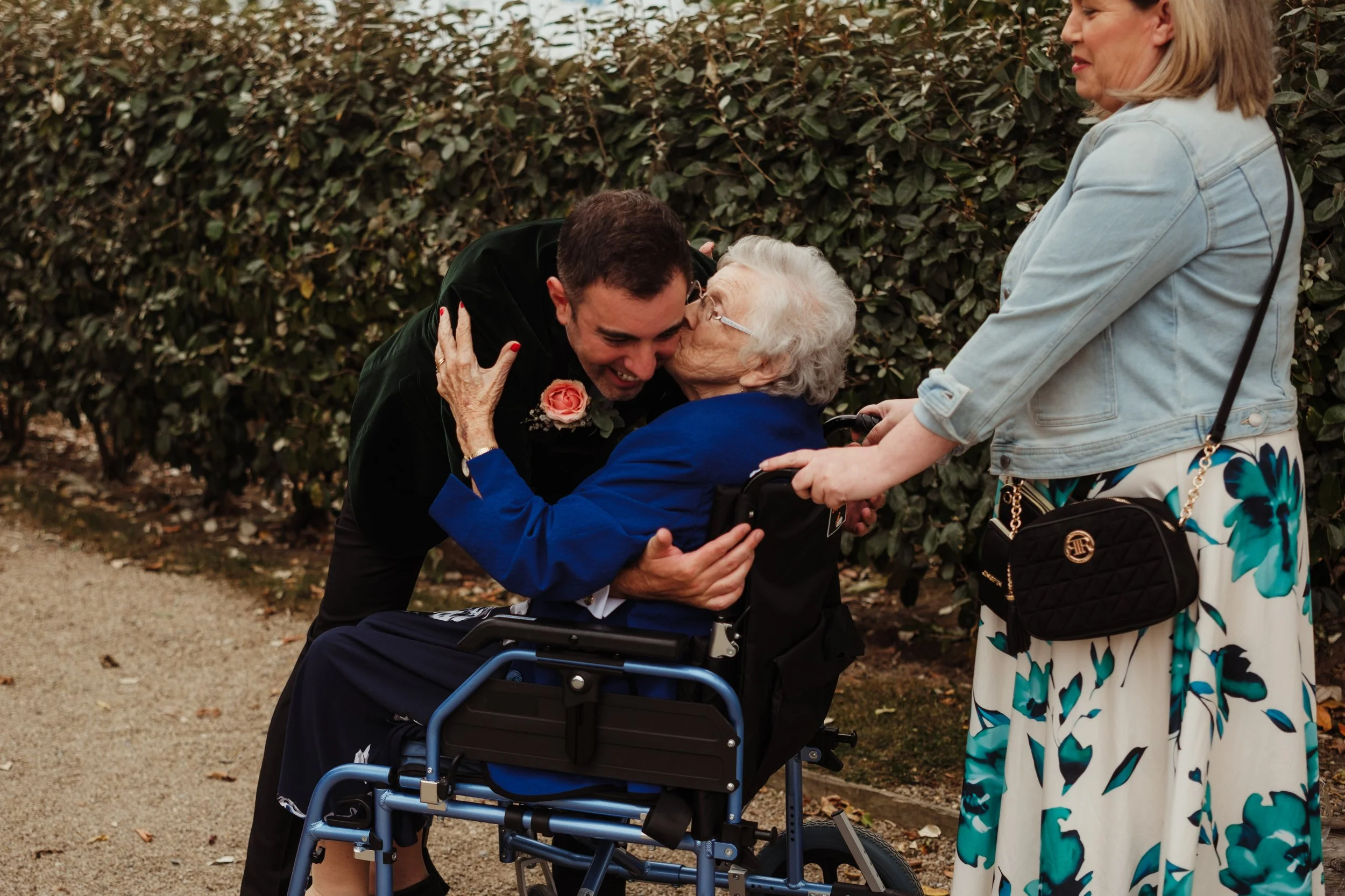 An elderly woman in a wheelchair is being kissed on the cheek by a young man in a black jacket. A woman in a light denim jacket and floral skirt is standing nearby, holding the wheelchair handle, outdoors in front of a hedge.