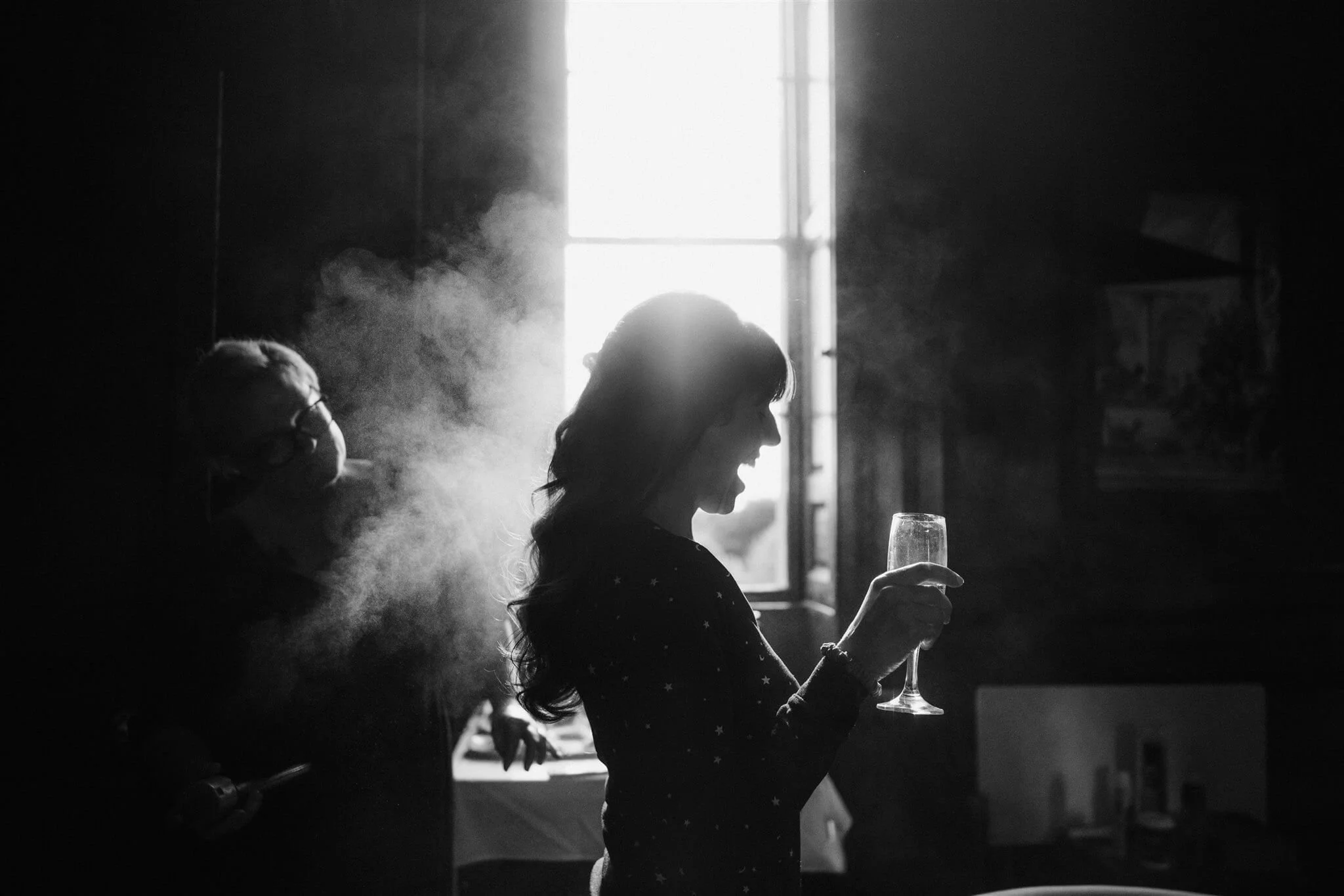 Silhouette of a woman holding a champagne glass in front of a bright window, with smoke or vapor around her, in a dimly lit room.