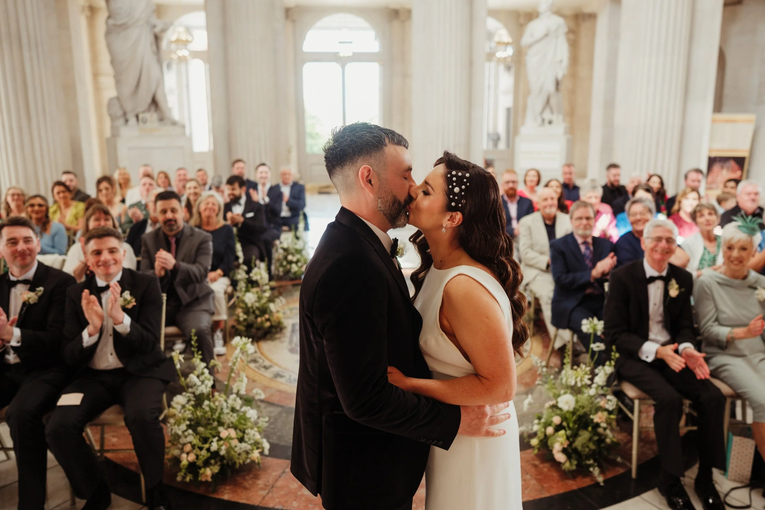 A bride and groom kiss during their wedding ceremony in a grand hall with large windows and statues, surrounded by seated guests applauding.