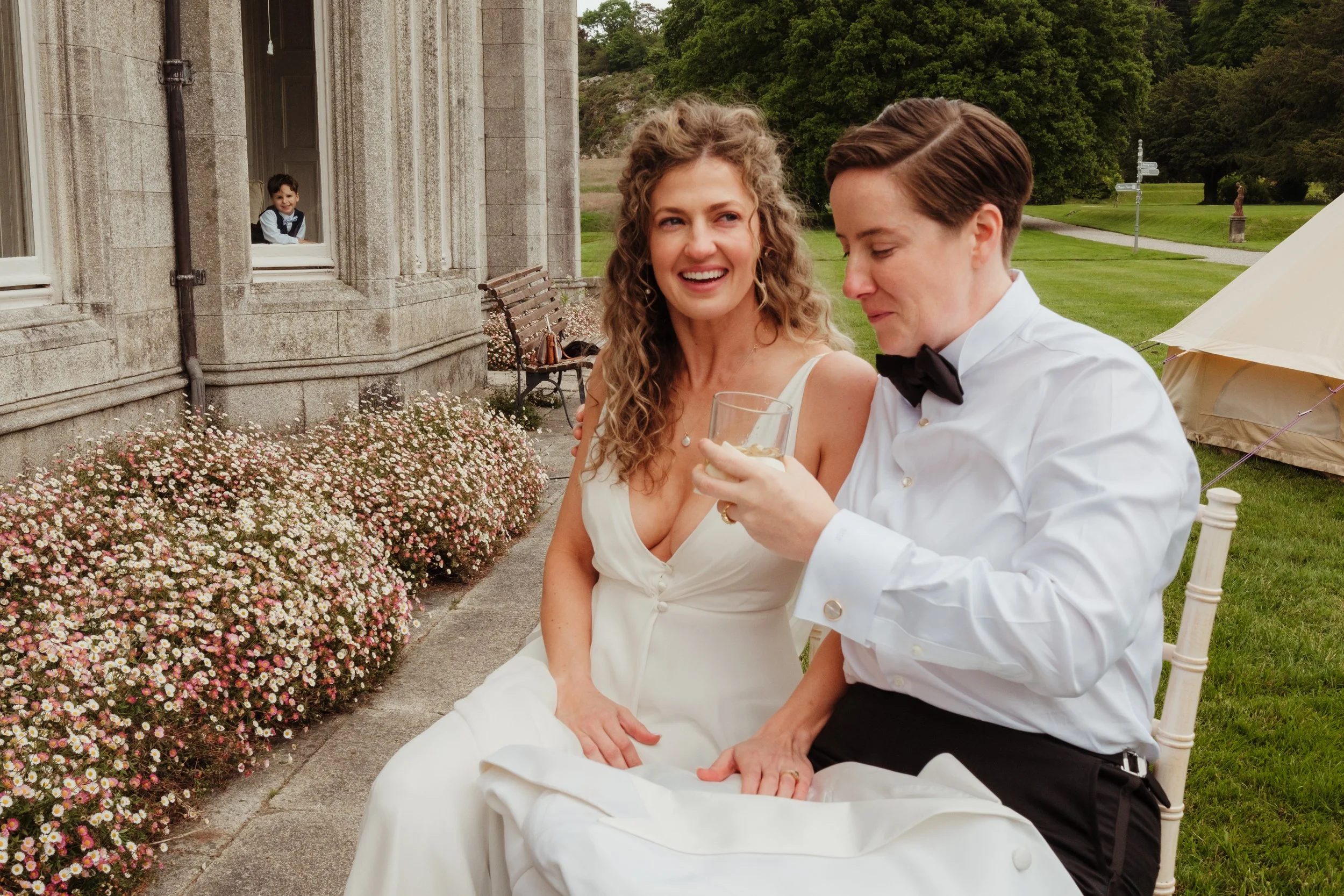 A woman in a white dress and a man in a tuxedo with a bow tie sitting outdoors, smiling and enjoying drinks. A young boy is looking out a window of a stone building in the background. There are pink and white flowers along the ground and green trees 