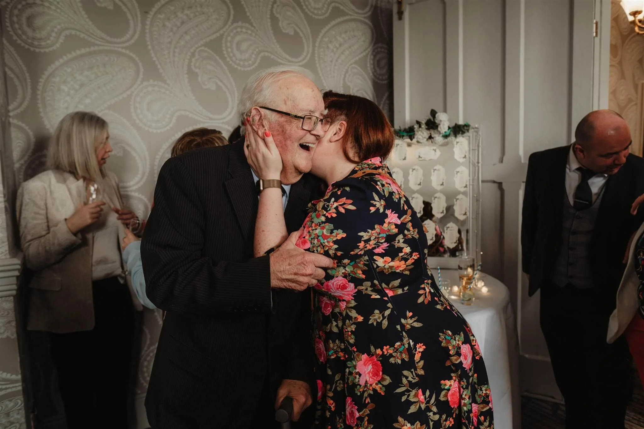 An elderly man with glasses and a woman with short red hair sharing a joyful kiss at a gathering. The man is smiling, and she is holding his face with both hands. In the background, other people are engaged in conversation and holding drinks. The set