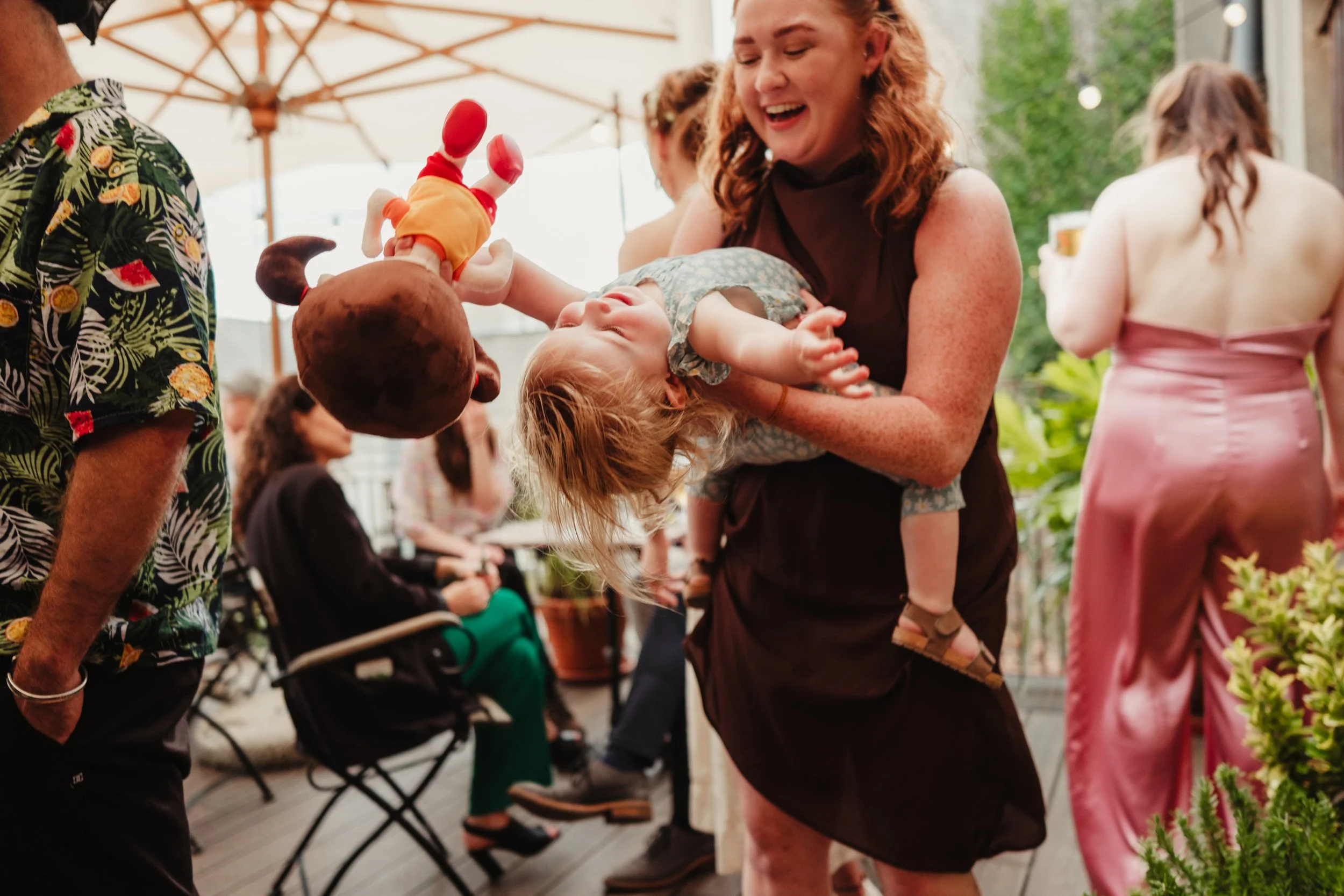 A woman holding a tilted child dressed in a patterned shirt, smiling and laughing at outdoor social gathering with others seated and standing around, some with drinks, under umbrellas and surrounded by green plants.
