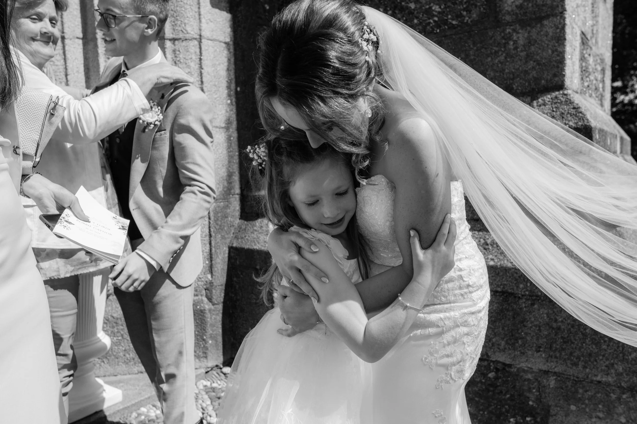 A bride in a wedding dress and veil hugging a young girl in a white dress at a wedding ceremony outside.