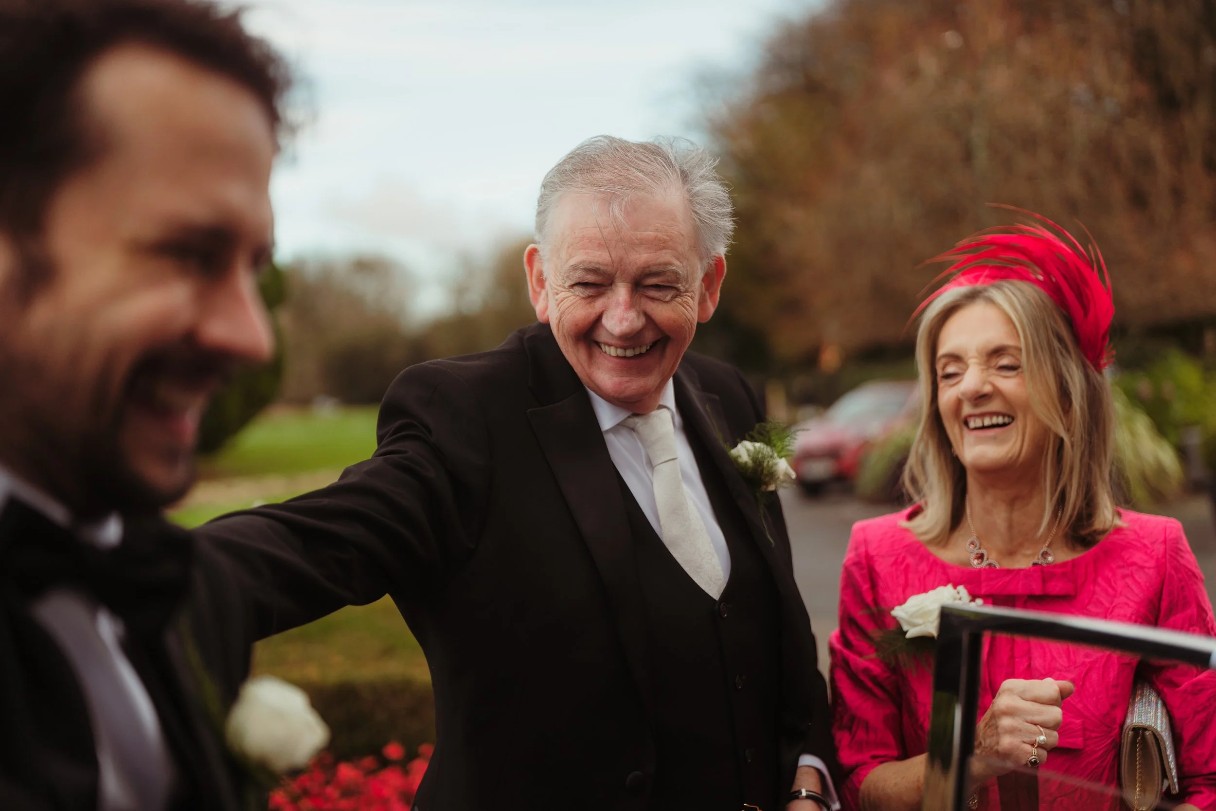 Three adults in formal attire smiling and laughing outdoors during a social event, with trees and parked cars in the background.
