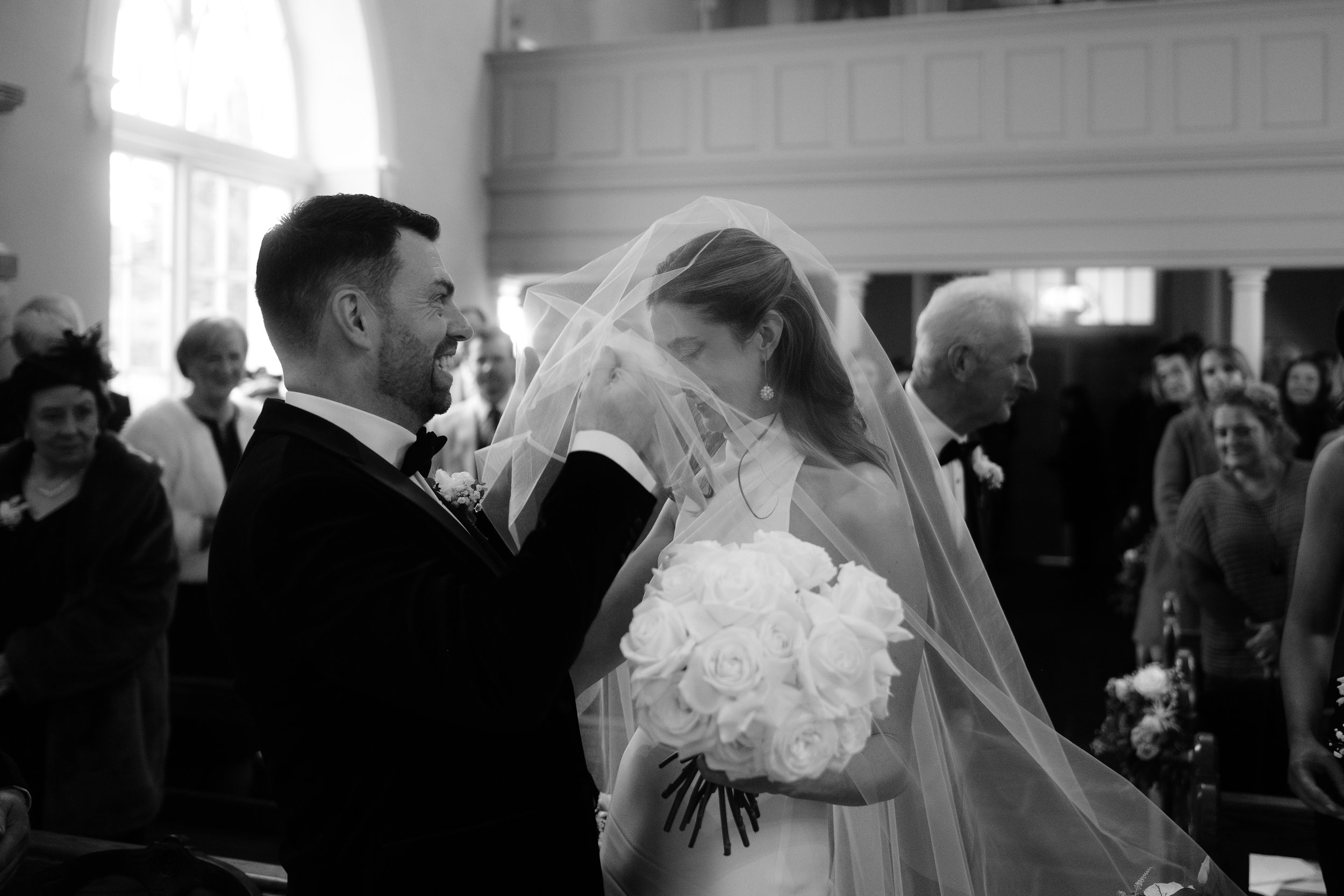 Black and white photo of a wedding ceremony inside a church, with a groom smiling and touching the bride's face, who is holding a bouquet of white roses and wearing a veil.
