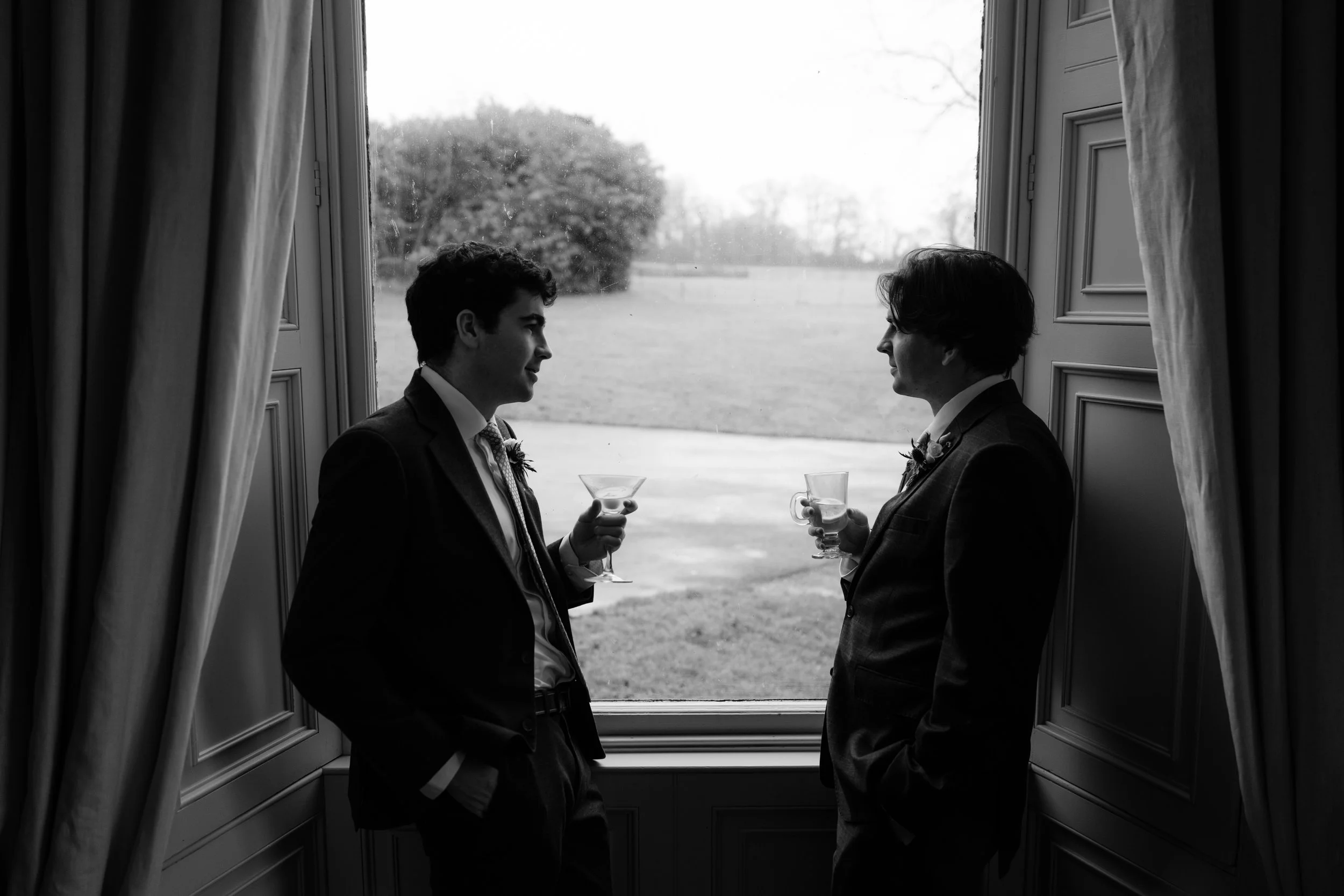 Two men in suits facing each other, holding cocktail glasses, standing by a large window with curtains, outdoors visible in the background.