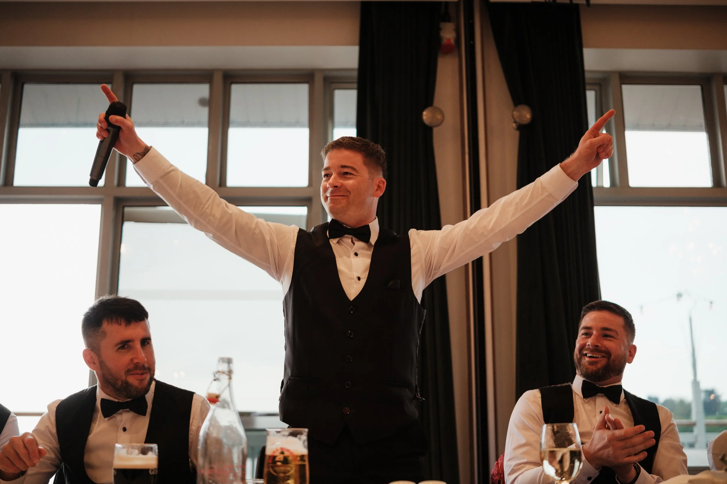 A man in formal attire, wearing a tuxedo vest and bowtie, holding a microphone and raising both arms, standing at a banquet table with two seated men in tuxedos looking at him and smiling.