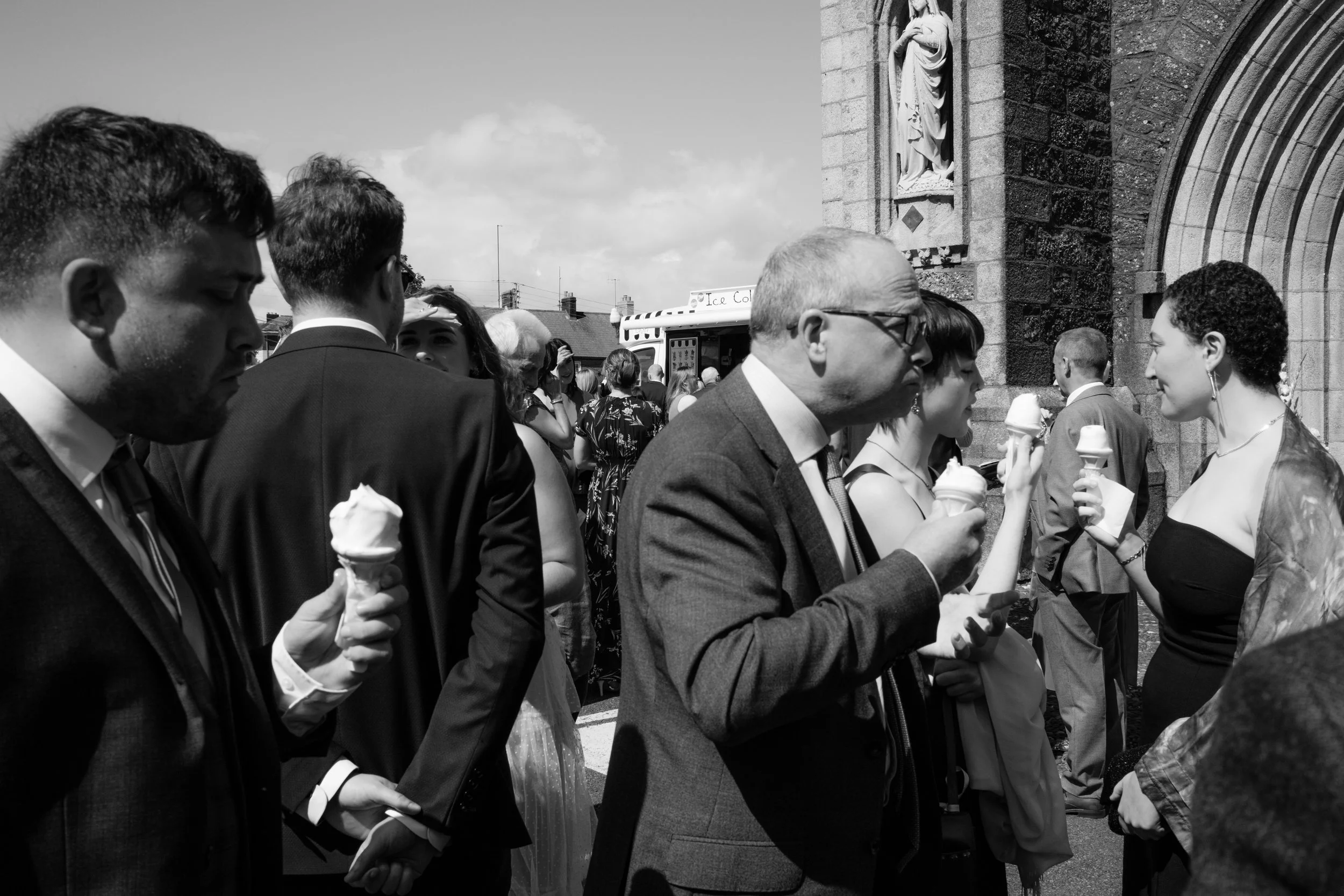 People in formal attire eat ice cream outside a stone church on a sunny day.