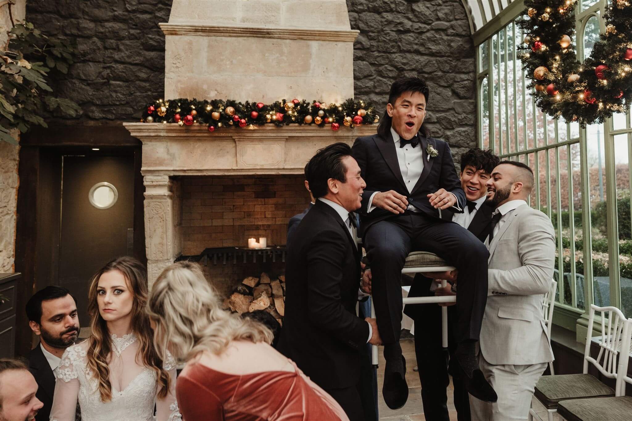 Group of men celebrations at a wedding, with one man being lifted in a chair in front of a fireplace decorated with Christmas ornaments, and a woman in a wedding dress nearby.
