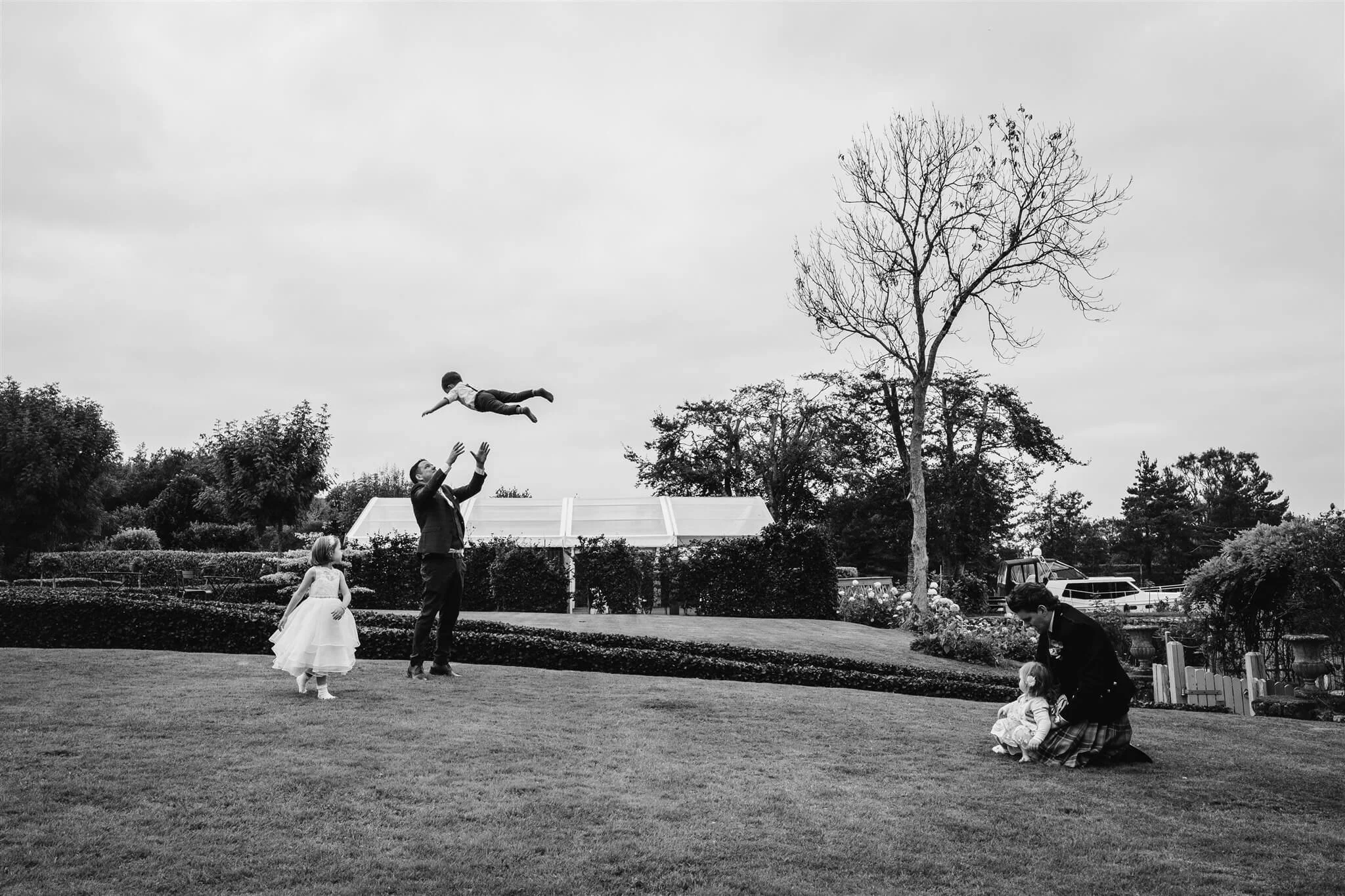 A man is tossing a boy into the air while two young girls watch and a man crouches down with a girl in a park with trees and bushes, in black and white.