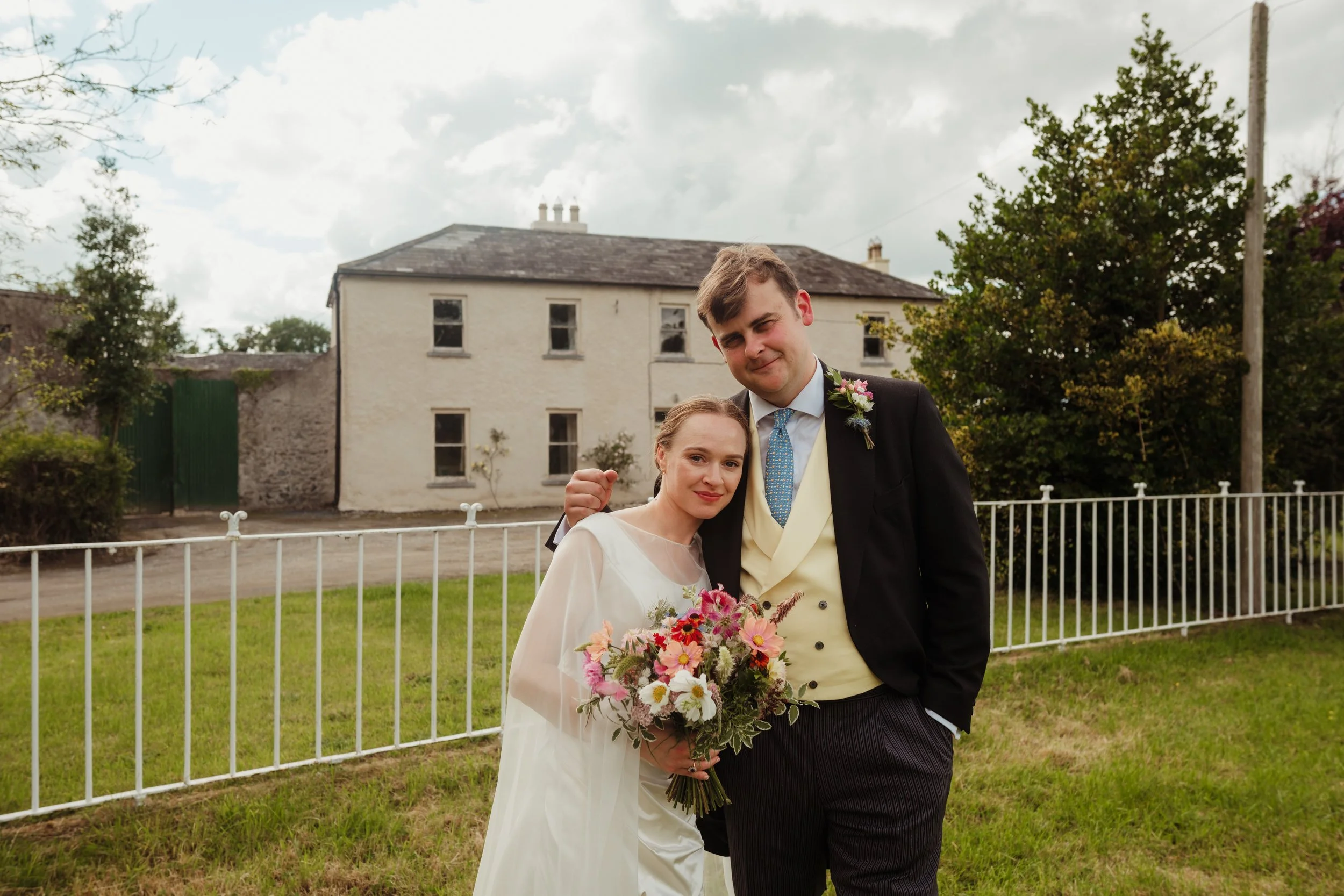 A bride and groom standing outdoors on a grassy lawn with a white fence behind them. The bride is holding a bouquet of pink and white flowers, and the groom has his arm around her, both smiling at the camera. The groom is wearing a black suit with a 