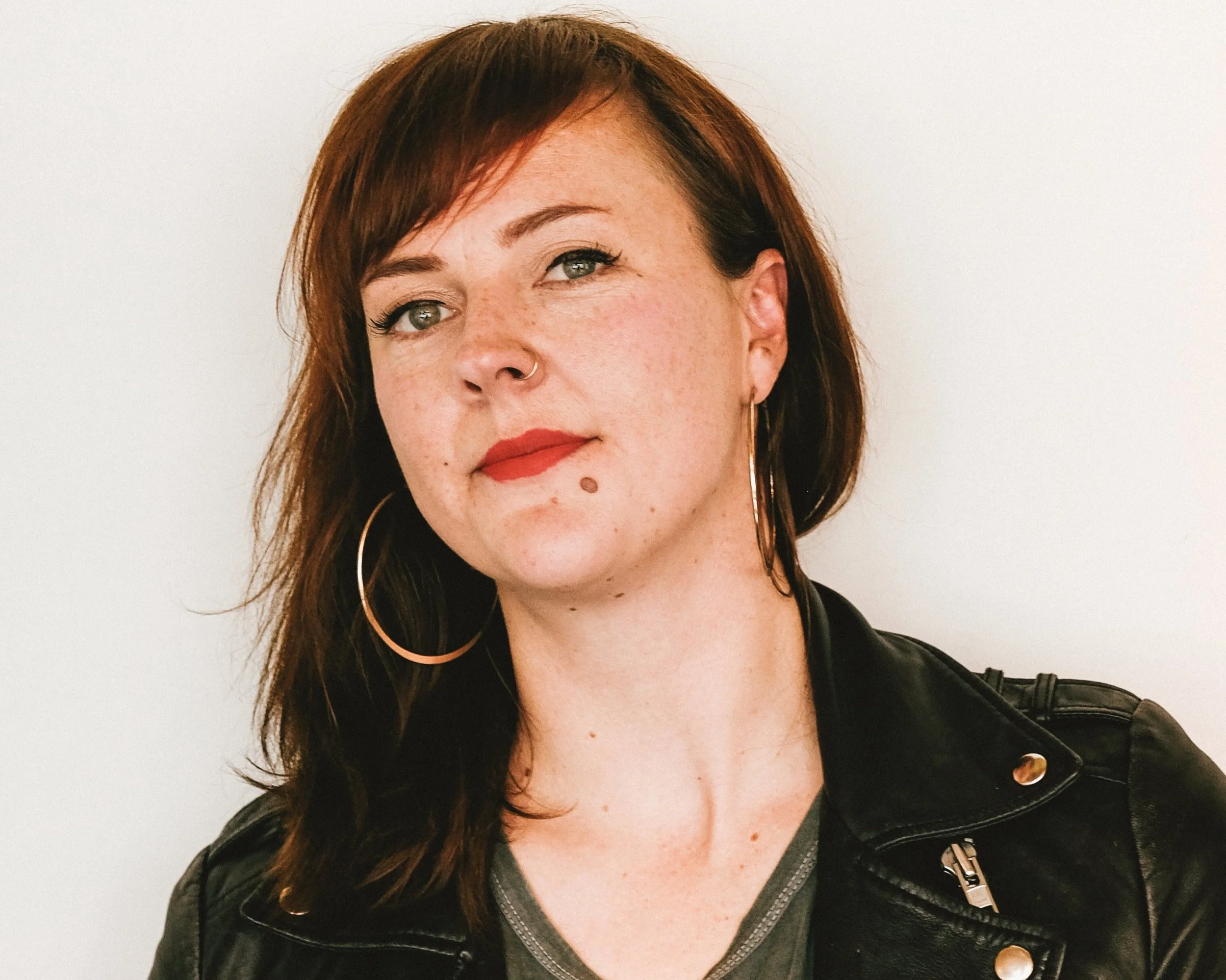 Emily is standing in front of a white wall. She has a black leather jacket on and has hooped earrings and red lipstick. She is looking at the camera.