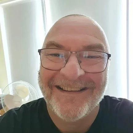 Malcolm is smiling looking at the camera with glasses on. He has a grey beard and a black t-shirt. In the background is a white fan.