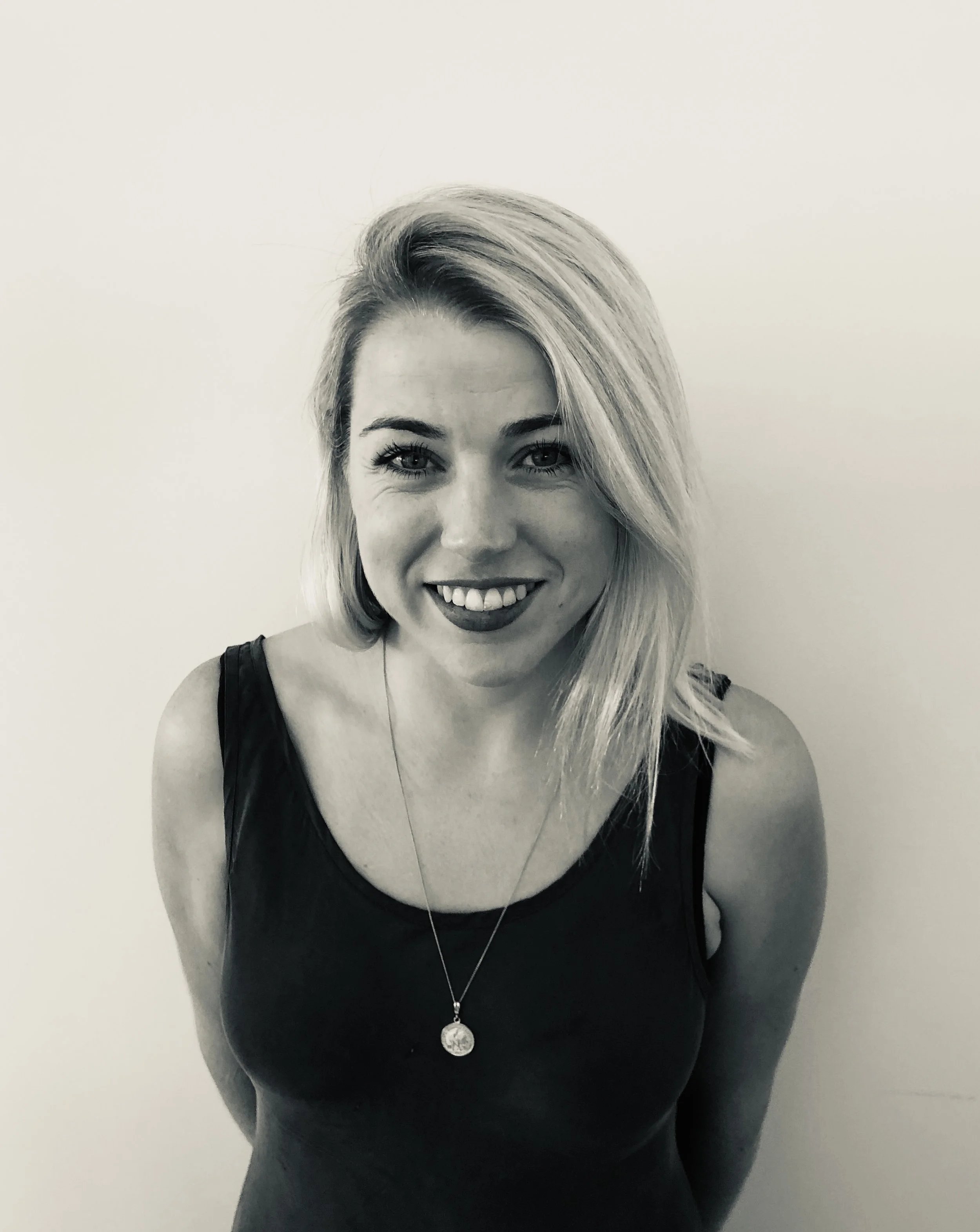 Katie has blonde hair which is sitting over one shoulder. She is wearing a black sleevless top and a necklace, she is standing against a white wall and is smiling directly into the camera. The photo is in black & white.