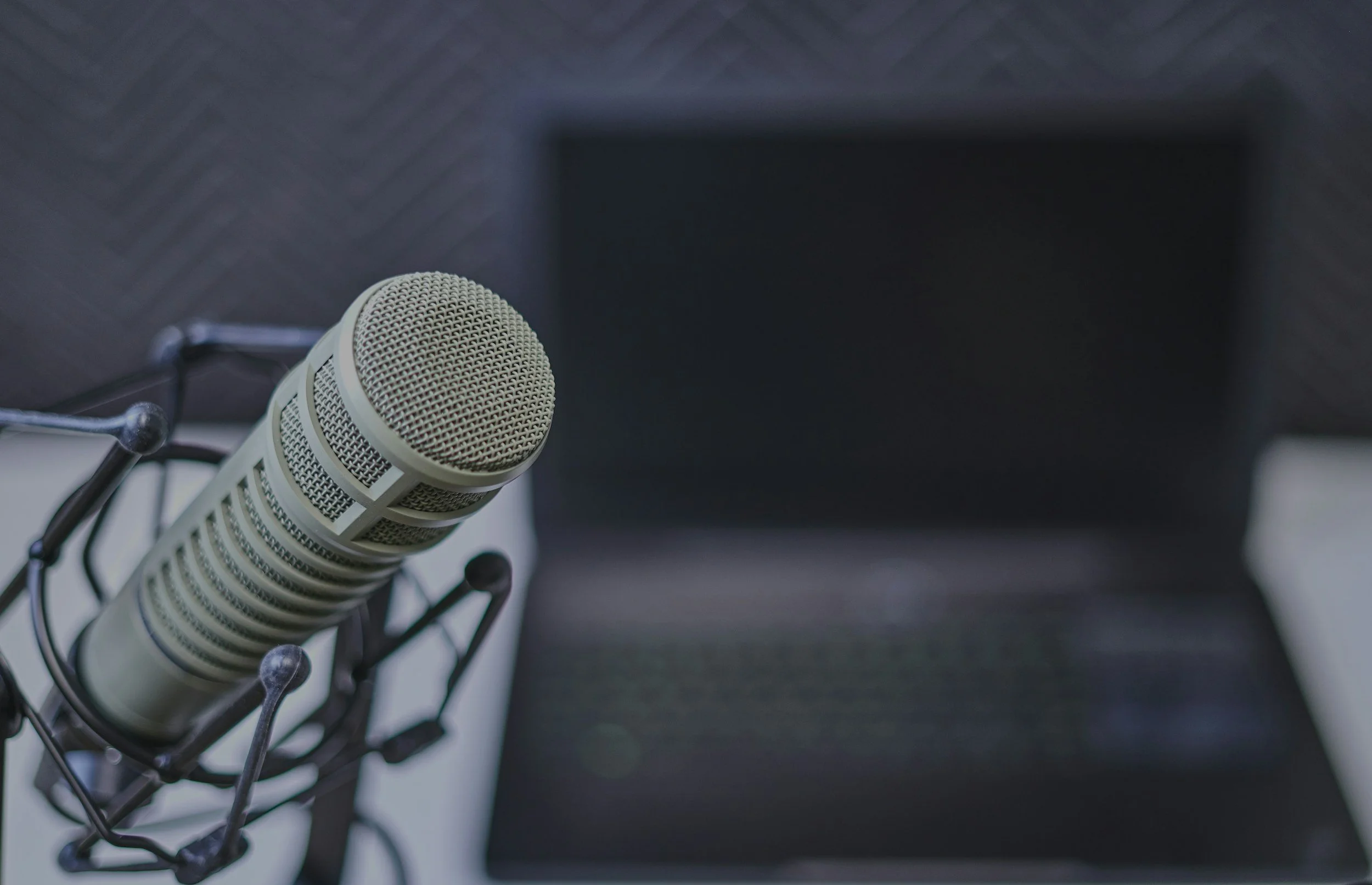 Close-up of a professional studio microphone mounted on a shock mount in a recording studio, with a blurred laptop in the background.