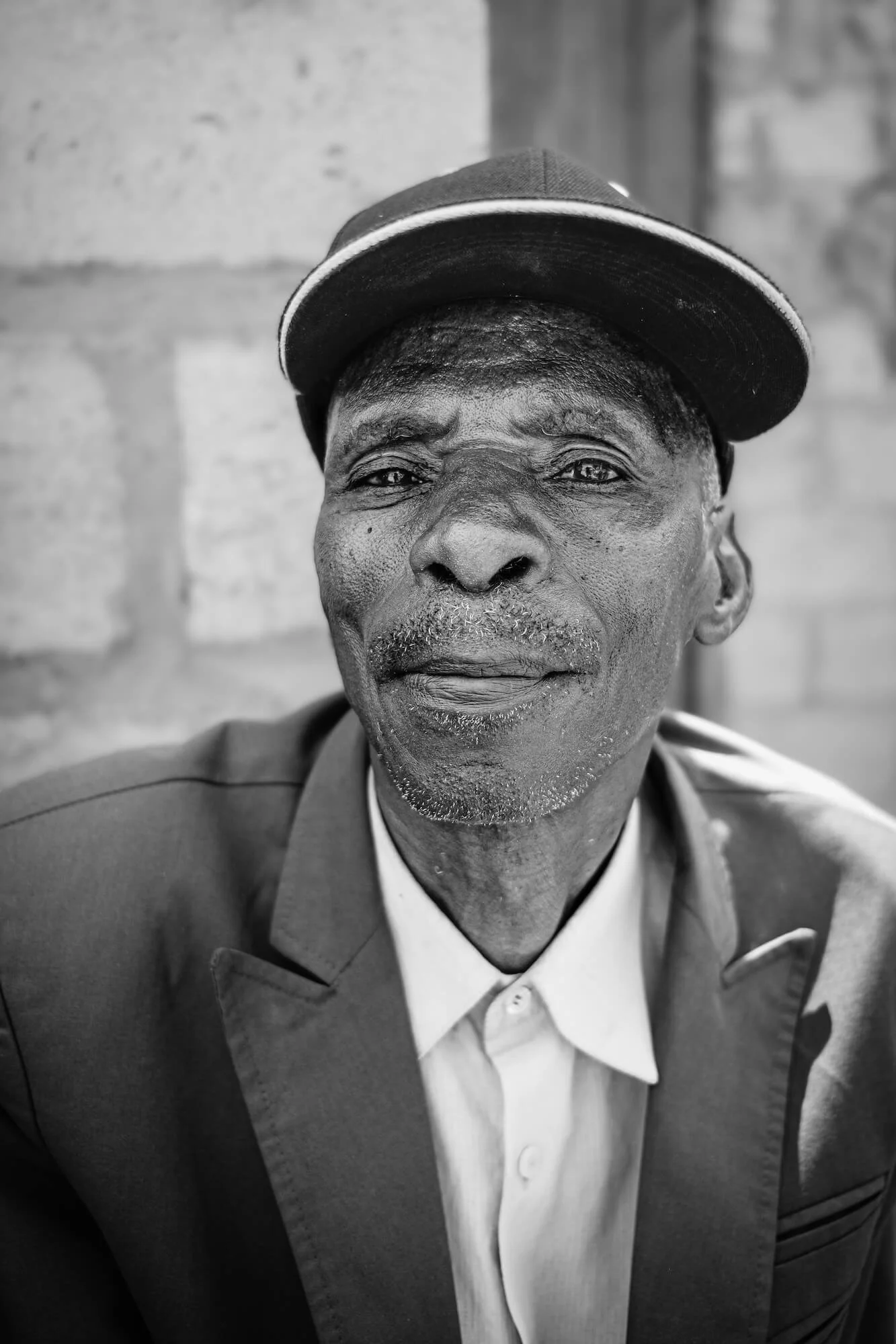 A black and white close-up photograph of an elderly Zambian man wearing a suit, shirt, and a cap, looking directly at the camera with a neutral expression.