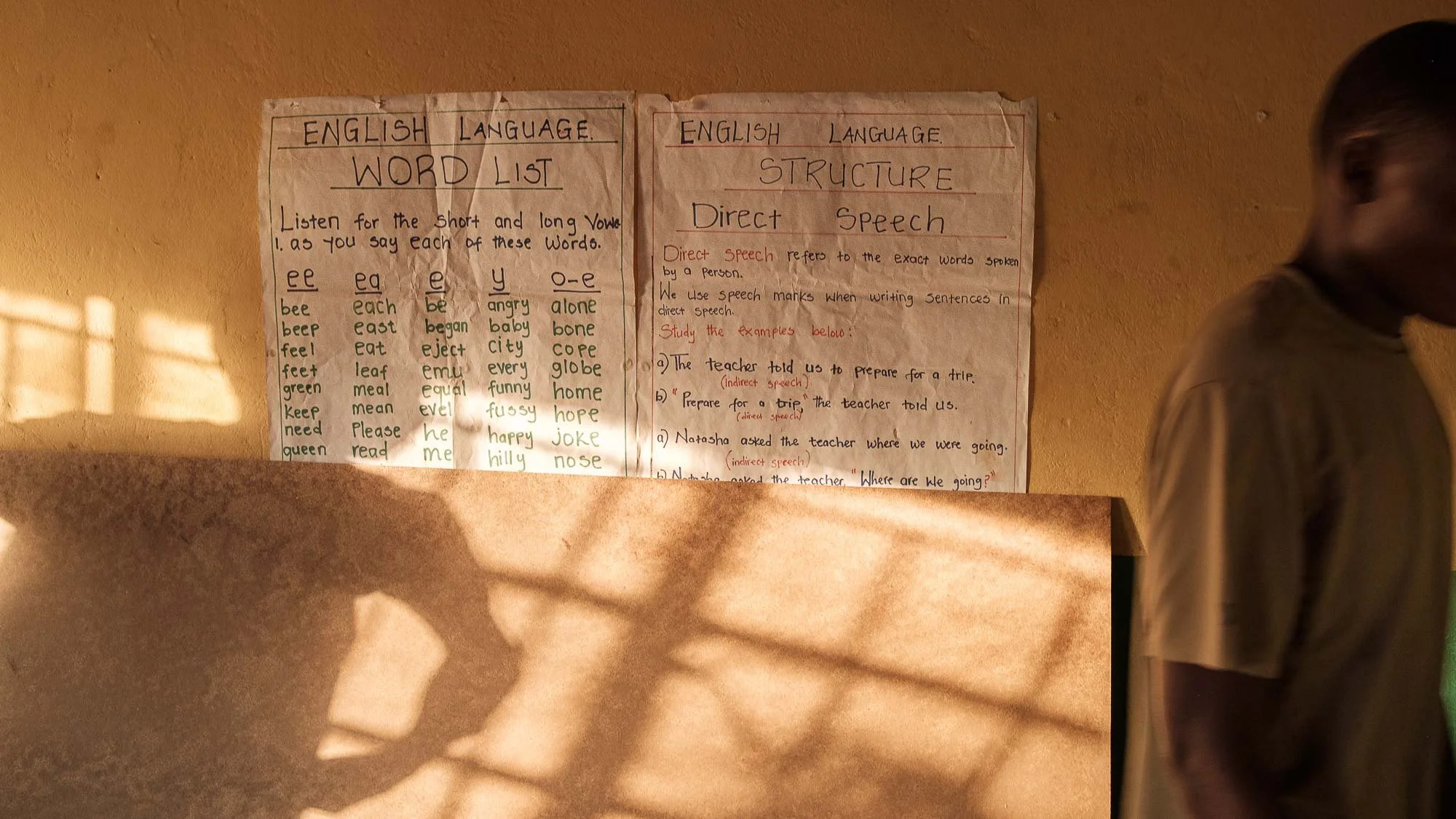 Educational posters on a wall with handwriting about English language word list and sentence structure, a person standing on the right side, and shadows cast on the wall.