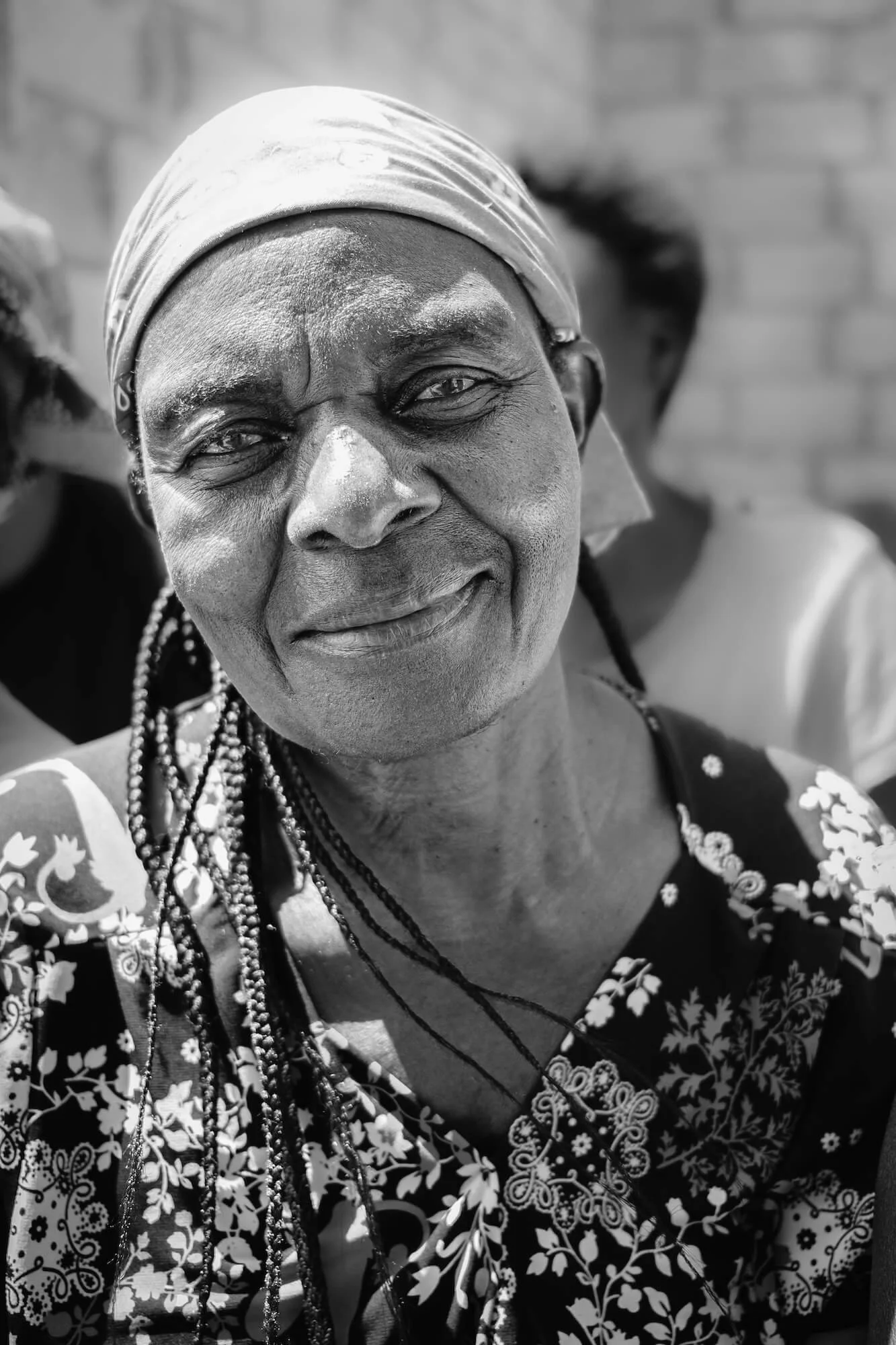 Black and white photo of a smiling Zambian woman wearing a patterned dress, headscarf, and necklaces, with a brick wall in the background.