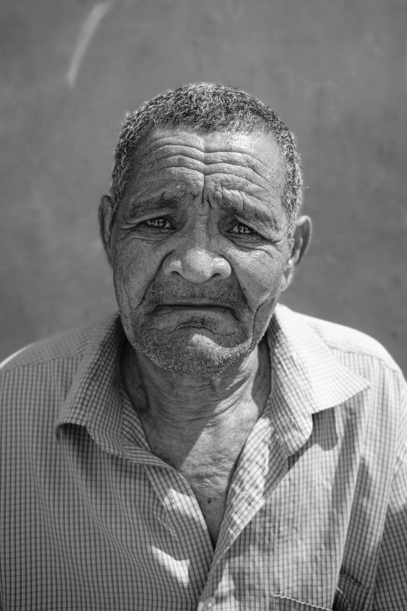 A black and white portrait of an elderly Zambian man with short, gray hair and deep facial wrinkles, wearing a checkered shirt, standing against a plain background.