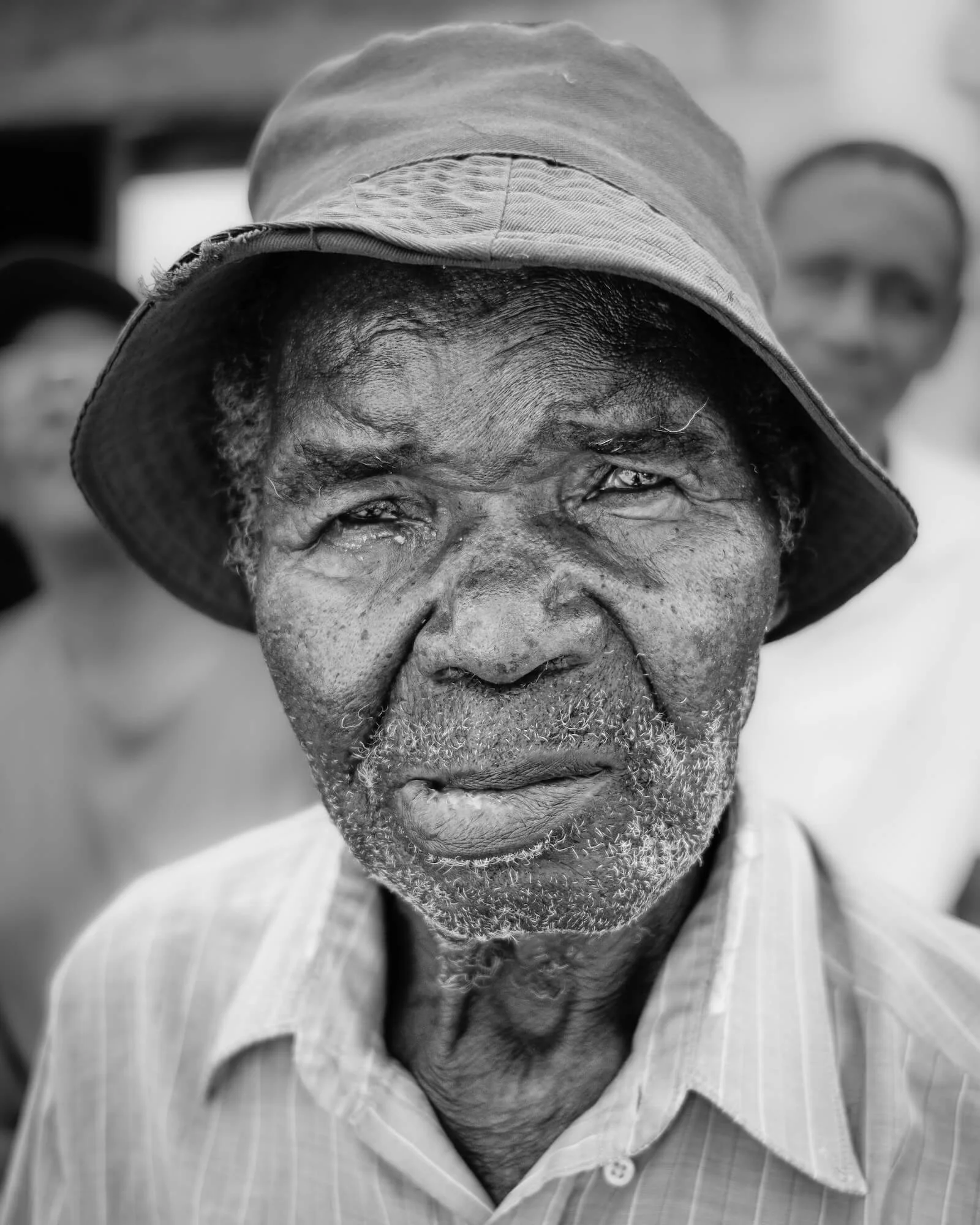 Close-up black-and-white portrait of an elderly Zambian man wearing a wide-brimmed hat and a striped collared shirt with a neutral expression, blurred people in the background.