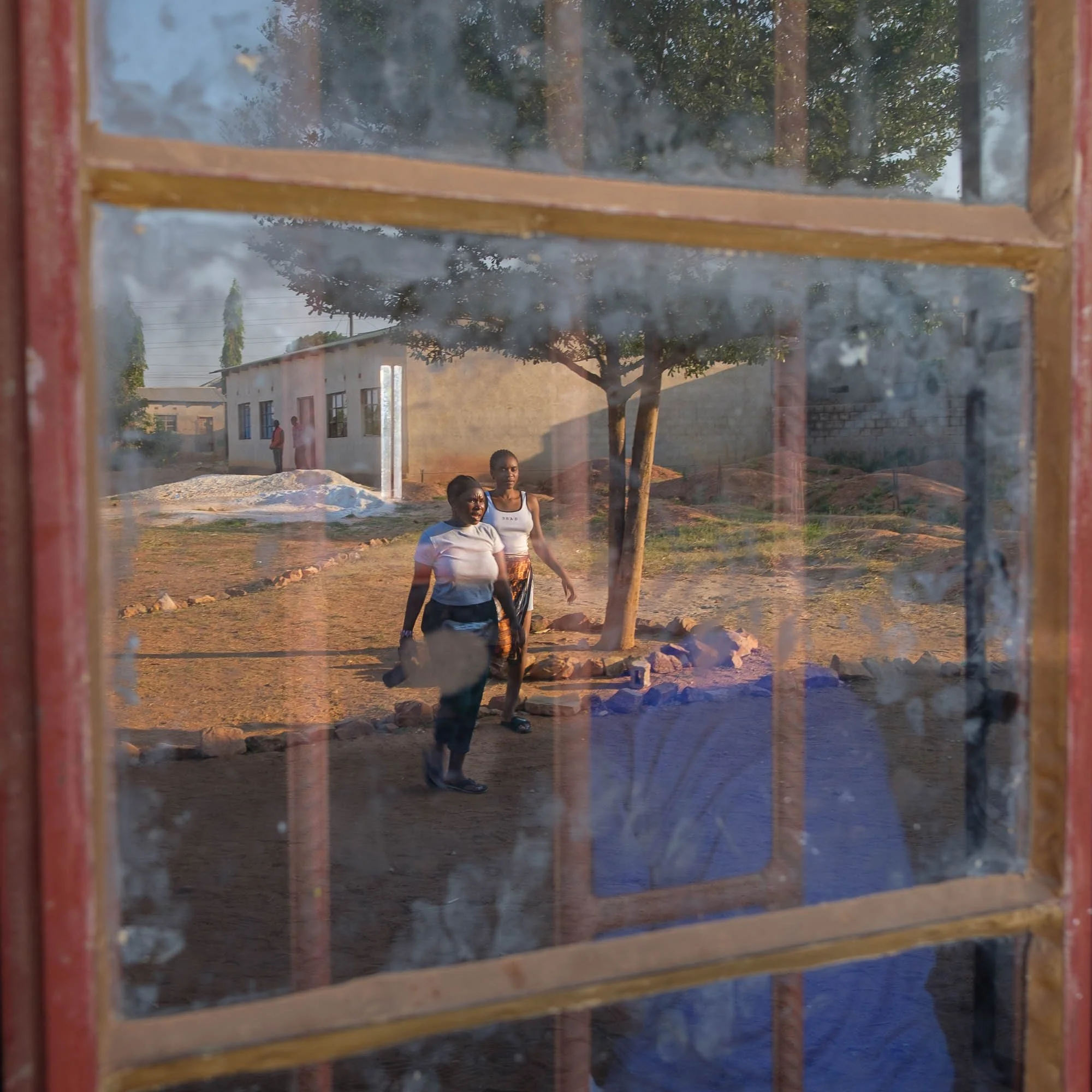 View through a window showing two women walking outside near a tree in a rural setting with a building in the background.