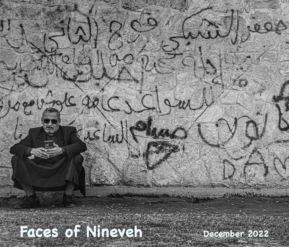 A man sitting against a wall with graffiti; he is wearing sunglasses, a dark coat, and pants with a pattern, and is looking at his phone. The wall behind him has handwritten Arabic script. Text at the bottom reads "Faces of Nineveh December 2022."