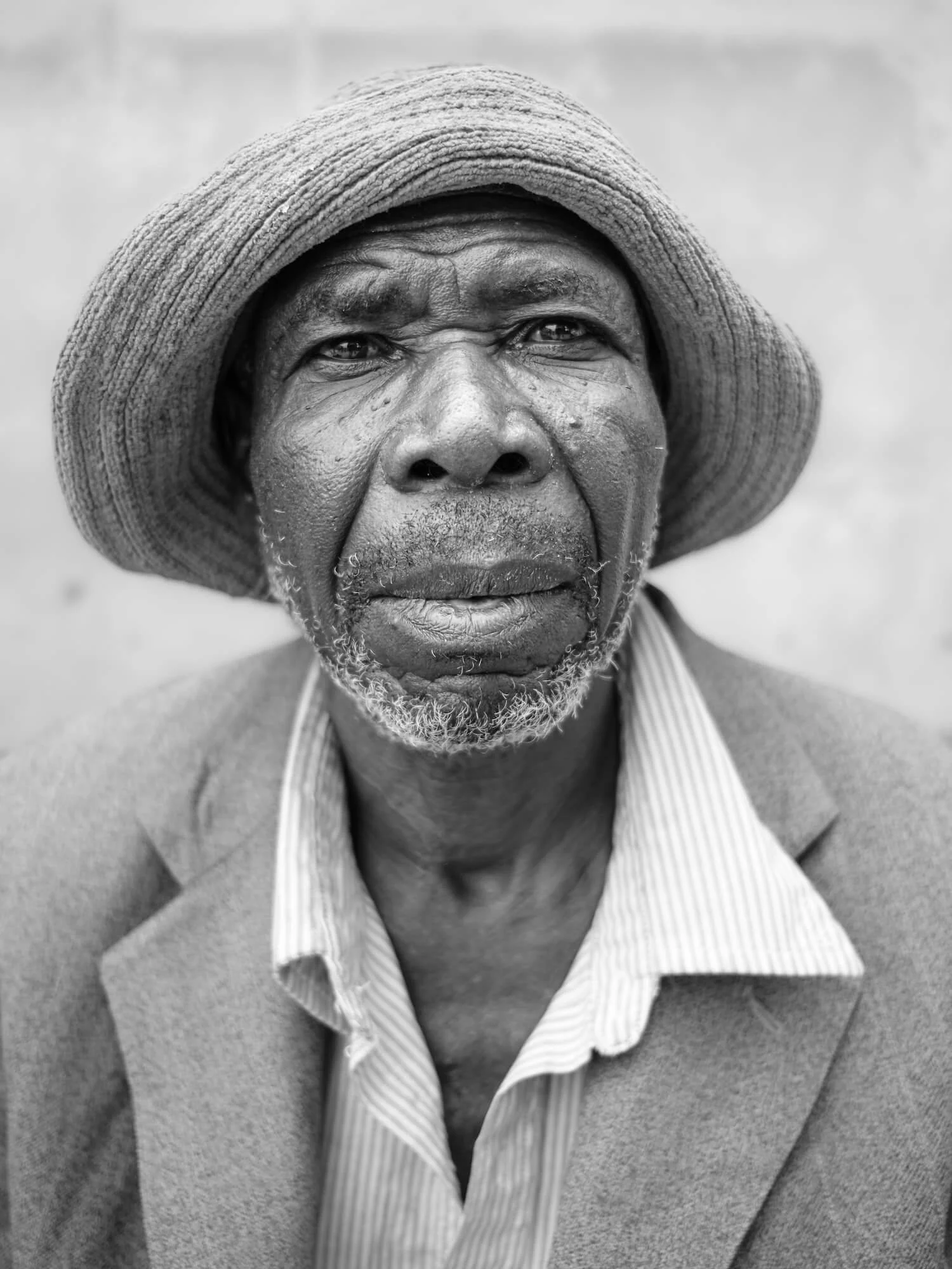 Close-up black and white portrait of an elderly Zambian man wearing a wide-brimmed hat, with textured skin and a beard, dressed in a suit jacket and collared shirt.