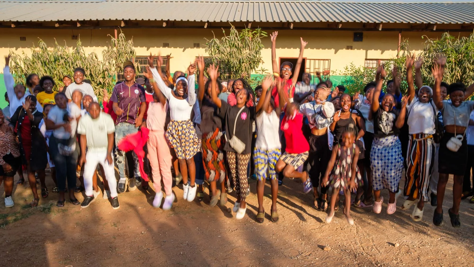 A large group of children and young adults are joyfully jumping and celebrating outdoors in front of a building with trees, in bright sunlight.