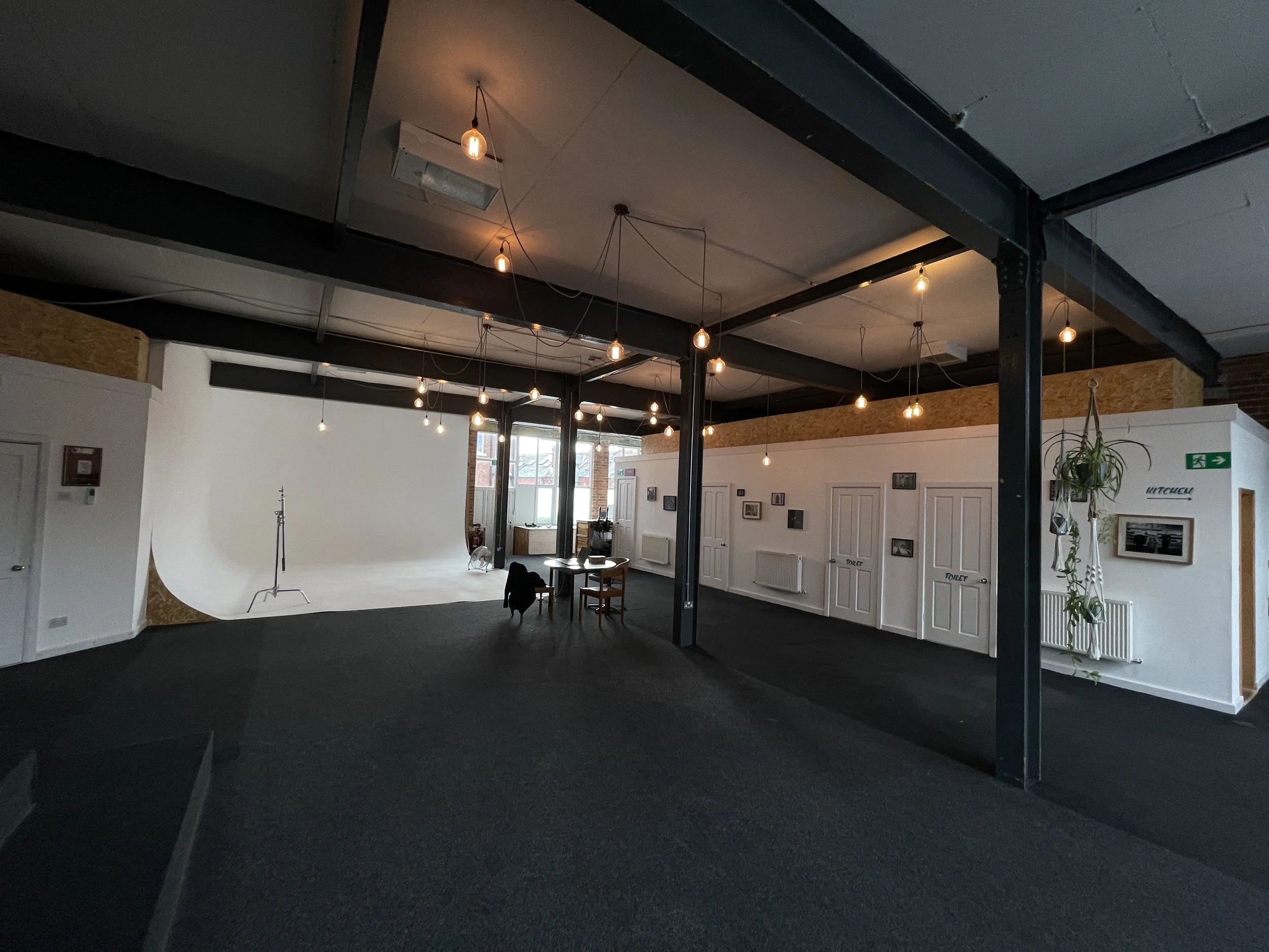 An interior space with a photo studio setup, including a white backdrop and lighting equipment, in a room with black flooring, white walls decorated with framed photos, hanging light bulbs, and a dining area with chairs and a table near large windows.