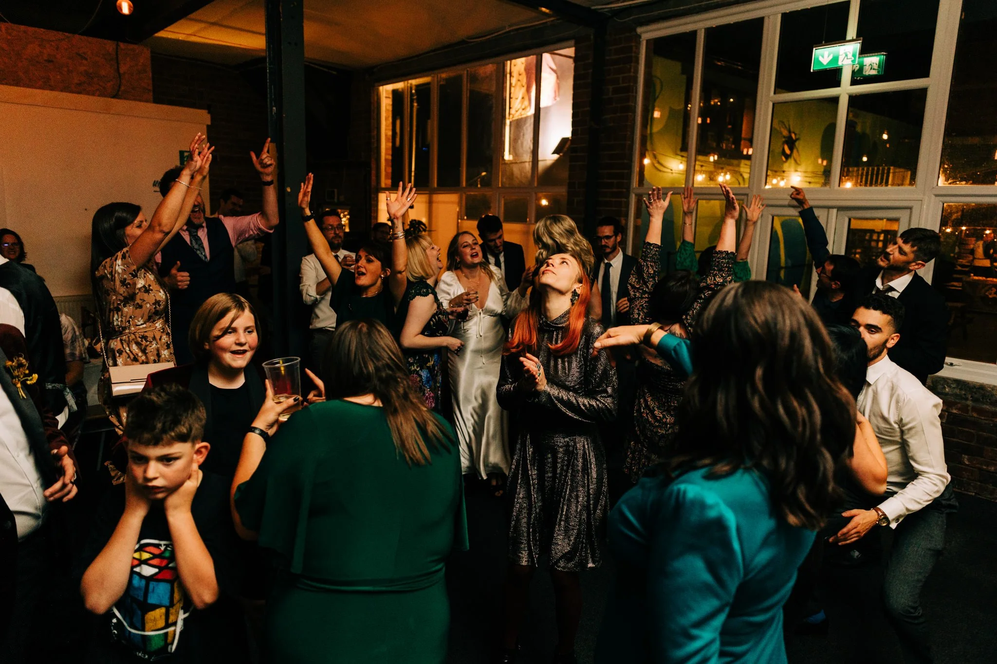 People dancing and socialising at a wedding with dim lighting and large windows.