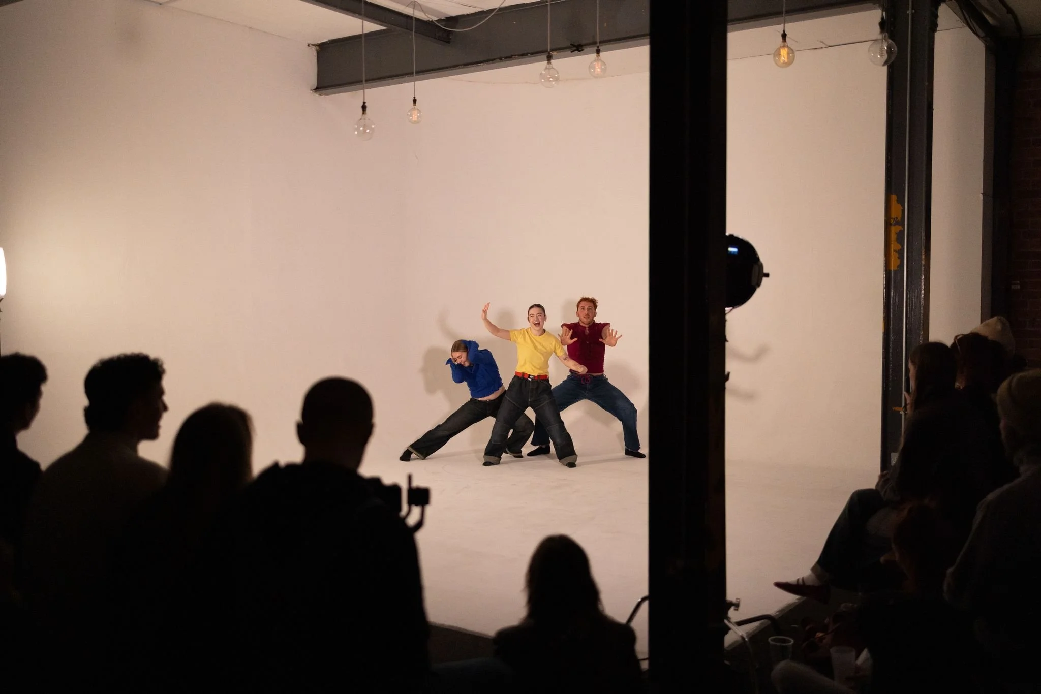 Group of three performers dancing in front of a white backdrop on a stage, with an audience watching in a darkened room.