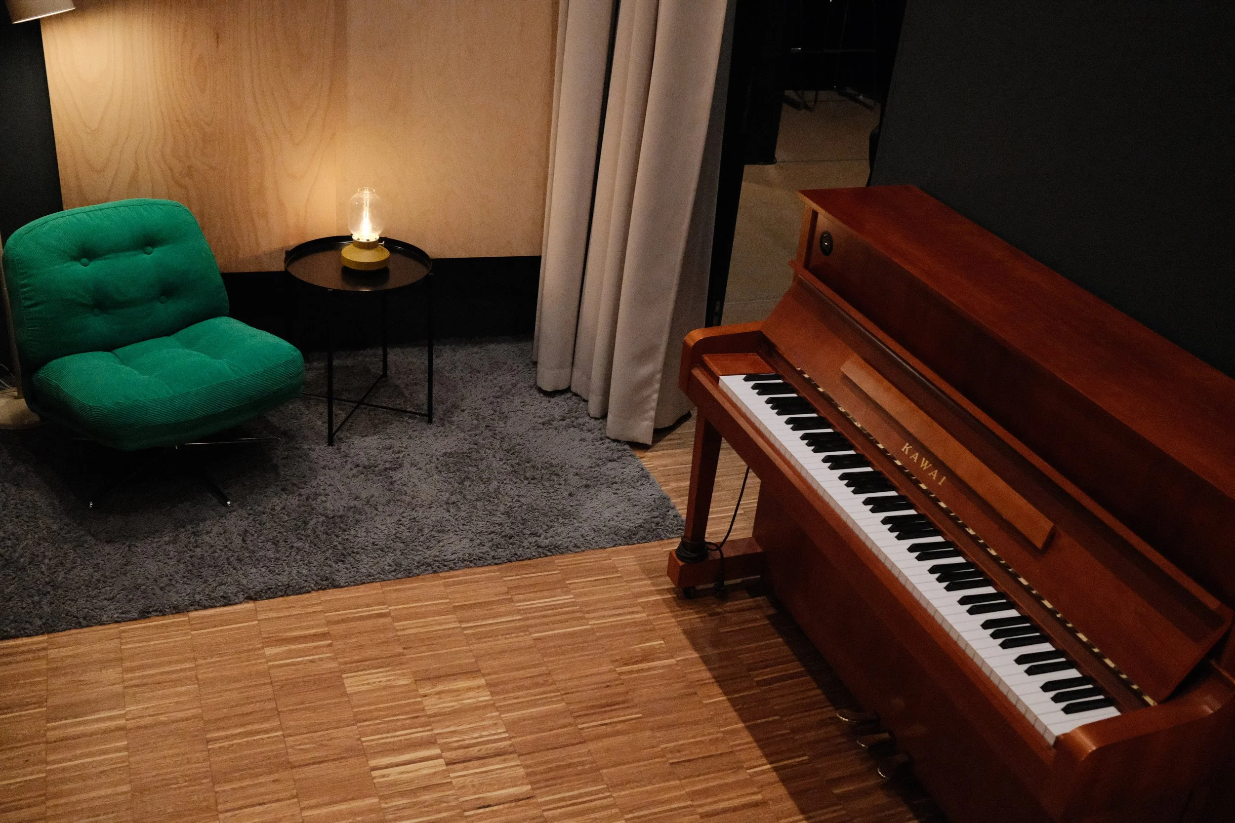 A cozy room corner with a green upholstered chair, a small round black side table with a decorative glass lamp, a wooden piano with black and white keys, a gray rug, beige curtains, and a wood-paneled wall.