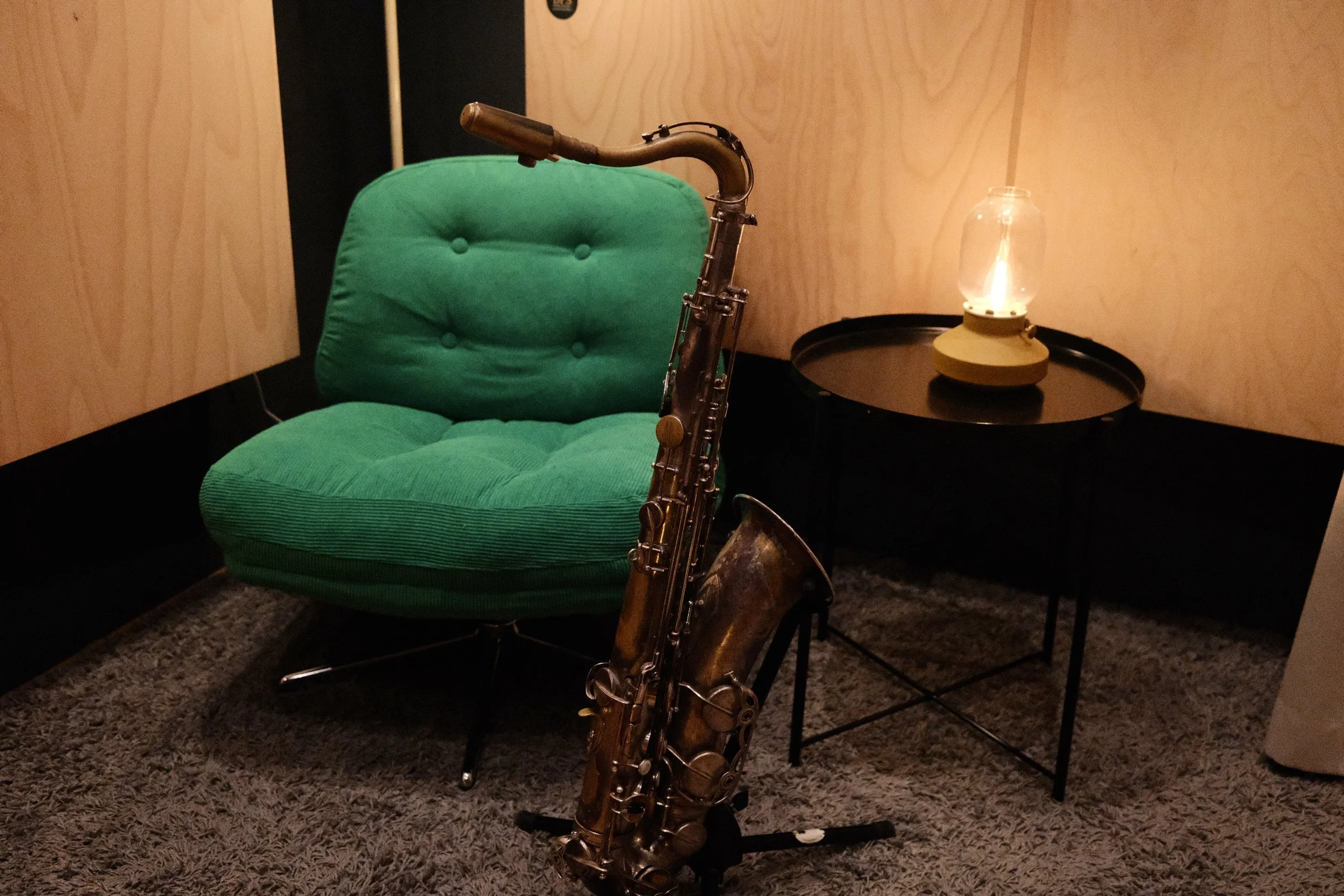A saxophone leaning against a black table with a yellow vintage oil lantern on it. In the background, there is a green cushioned chair with a wooden wall behind it.