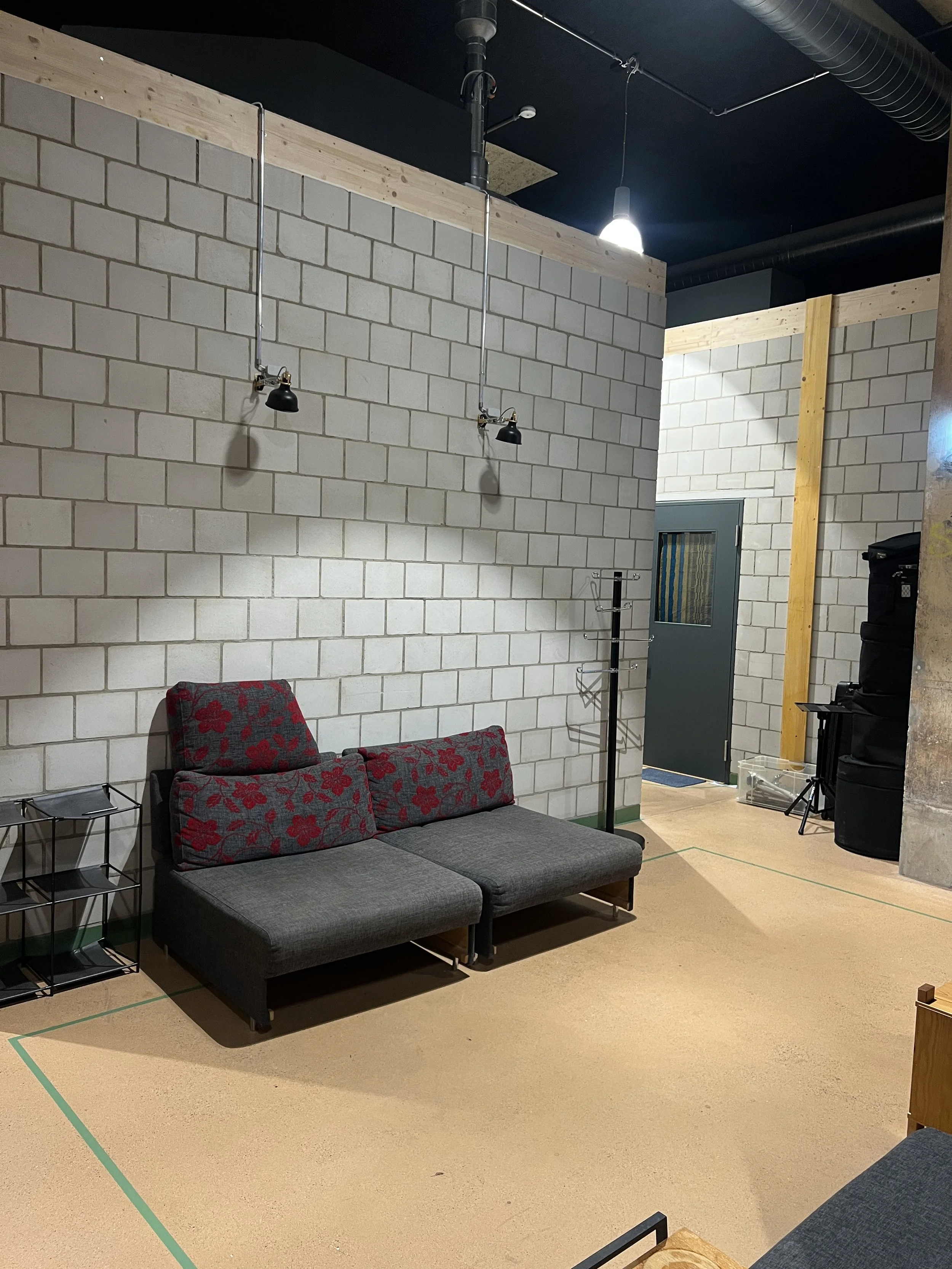 View of a waiting room with brick walls, a gray door, a gray couch with floral cushions, a small black side table, and hanging lamps. The ceiling has exposed ductwork and pipes, with a black ceiling.