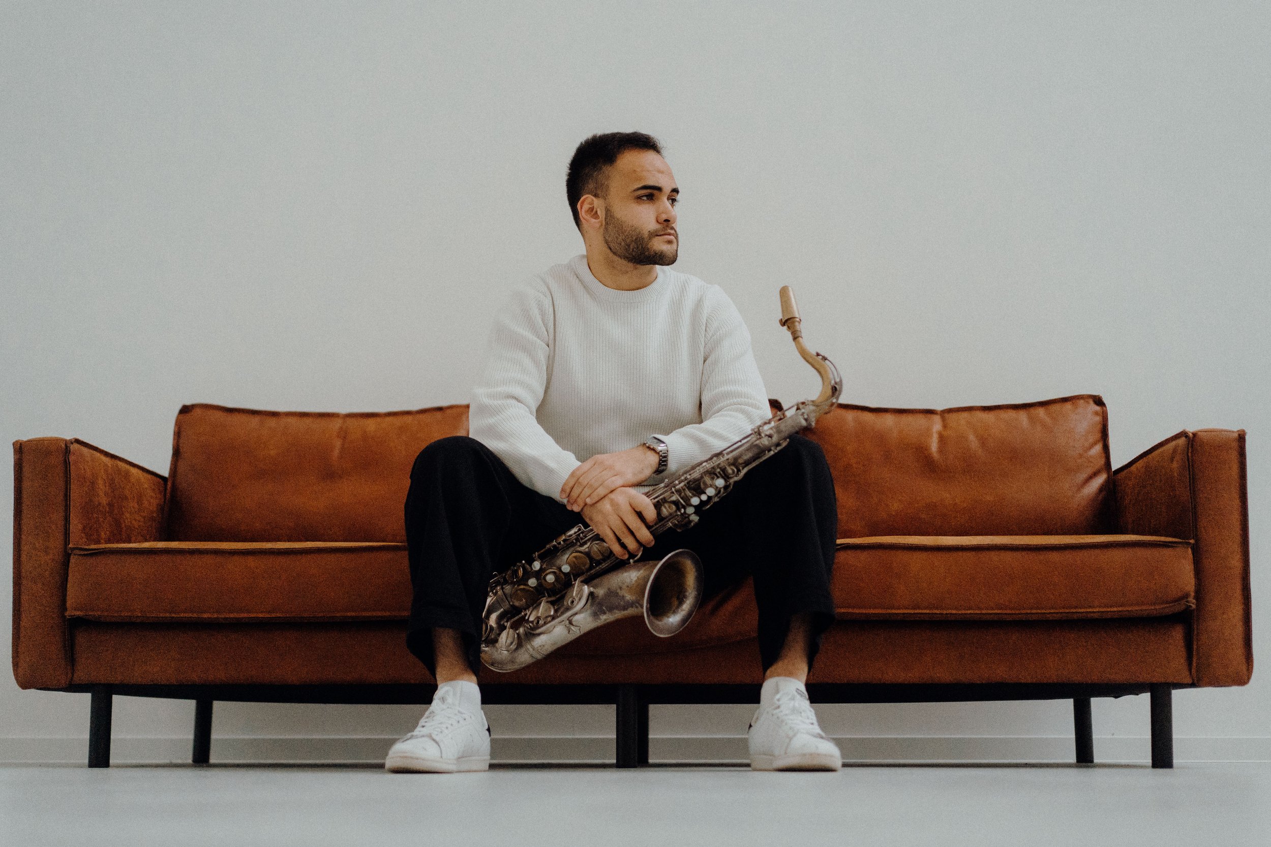 A teacher sitting on a brown leather sofa, holding a saxophone, wearing a white sweater, black pants, and white sneakers, in front of a plain white wall.