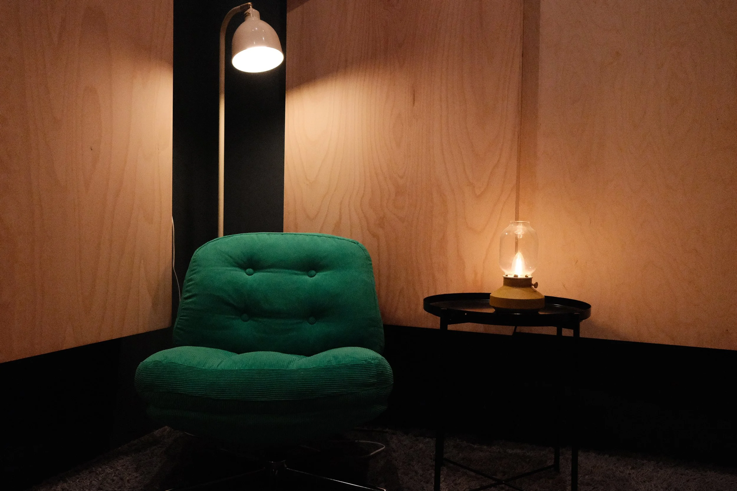 A cozy interior scene with a green chair, a black side table, a lamp, and a wooden wall background.