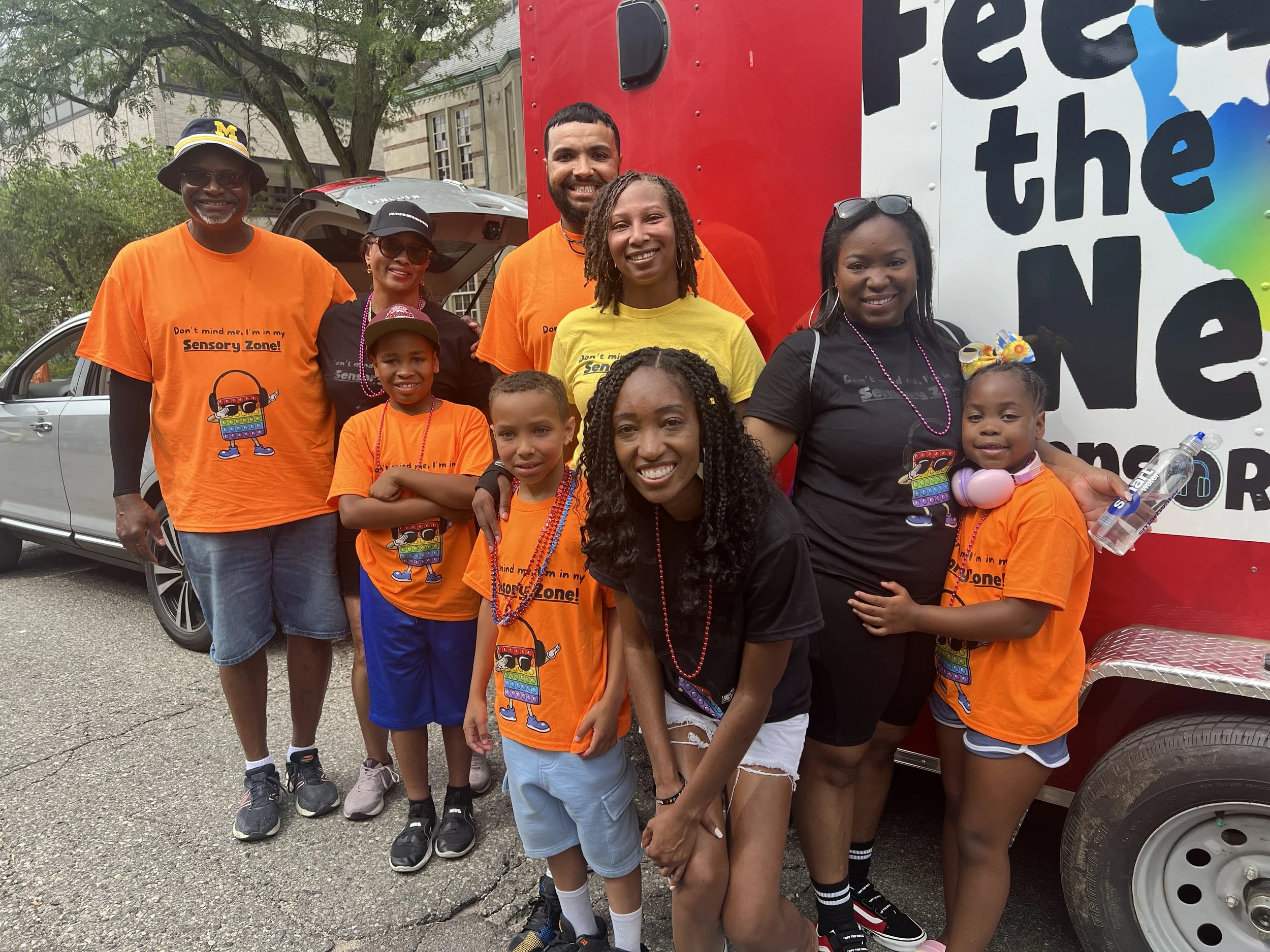 Group of diverse people, including children and adults, posing outdoors in front of a truck with a colorful banner, wearing matching orange and black shirts with a cartoon character, participating in a community event.