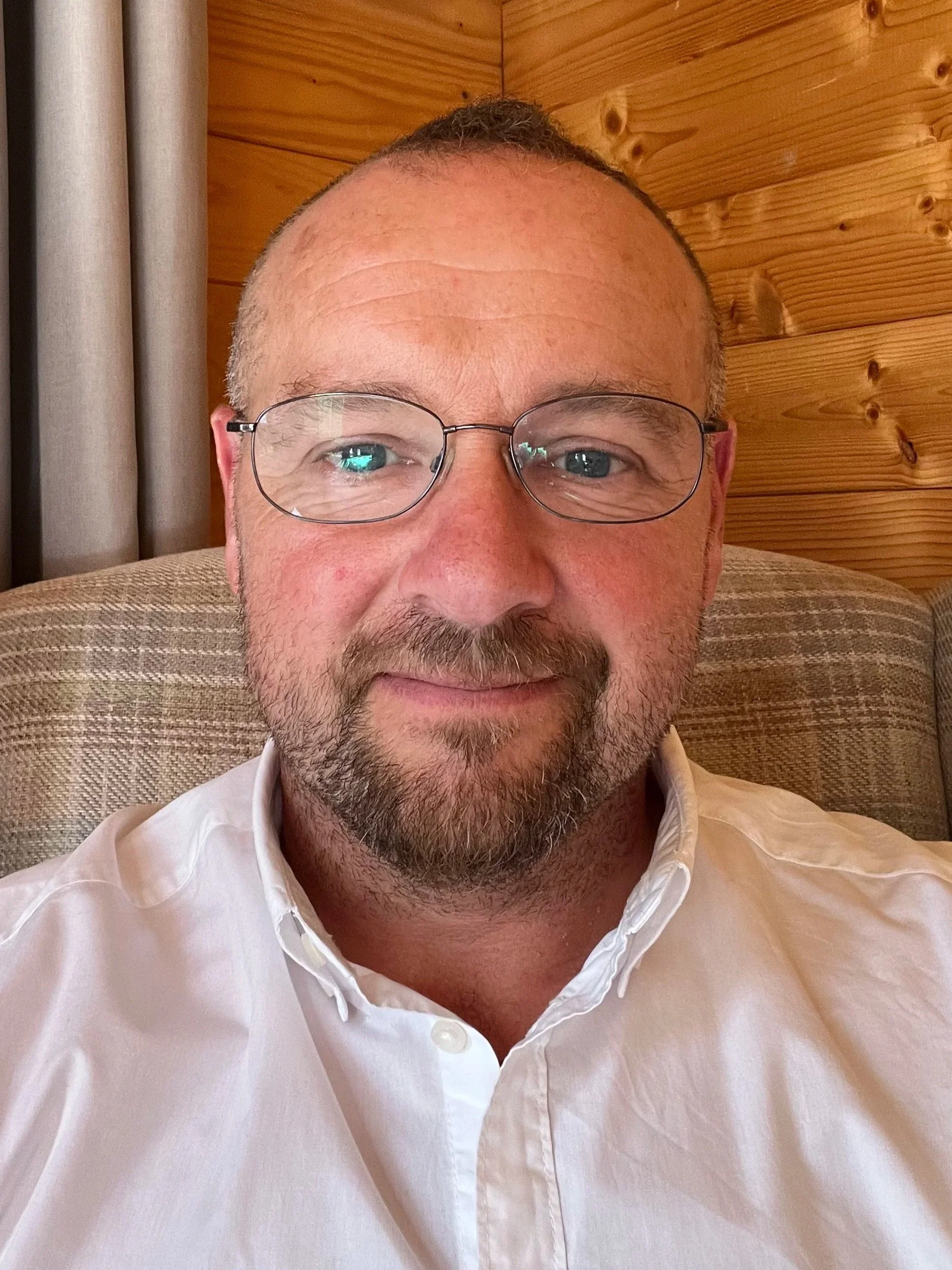A smiling middle-aged man with glasses and a beard, wearing a white button-up shirt, sitting on a beige couch with a wooden wall background.
