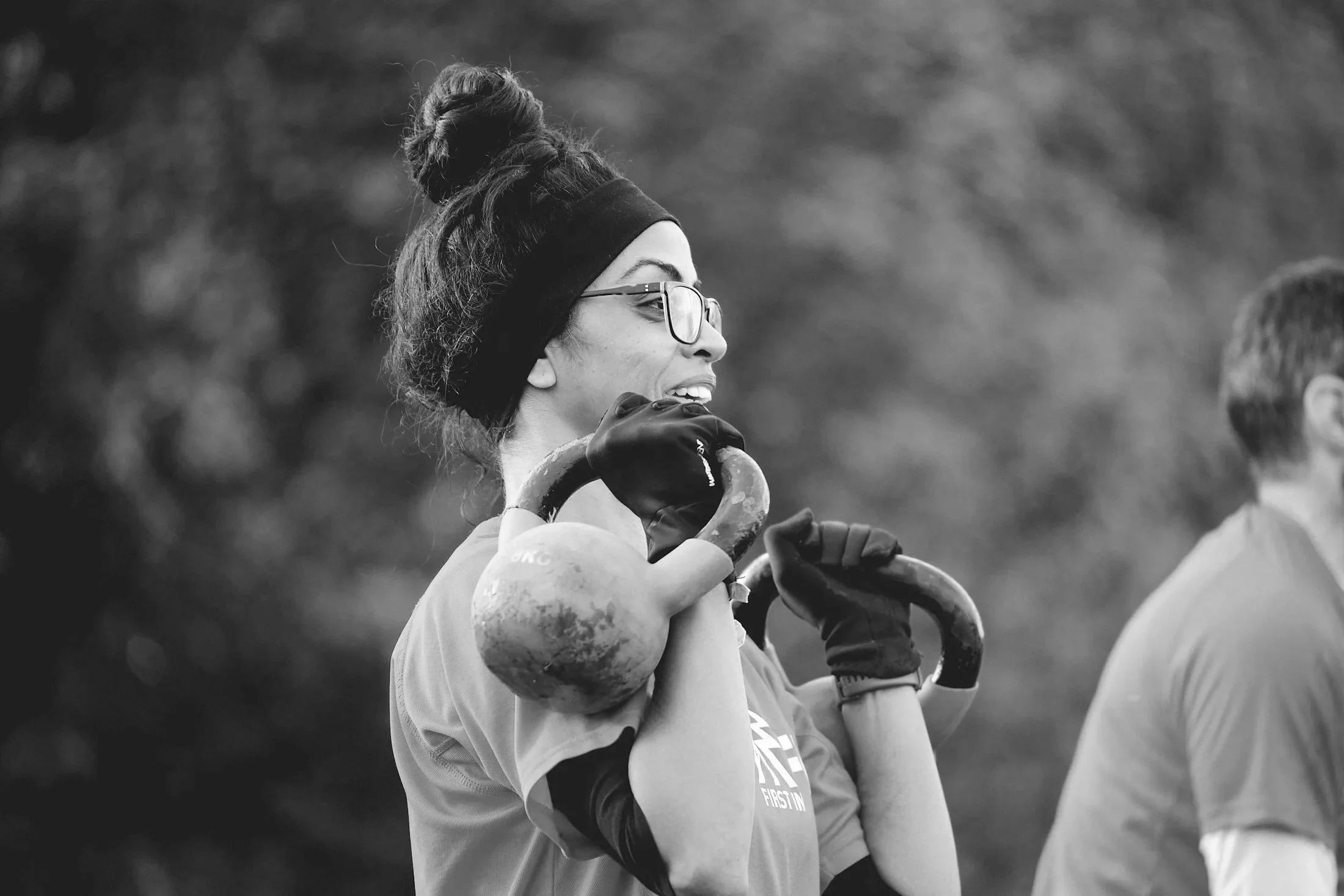 A woman with glasses, a headband, and gloves, holding a kettlebell on her shoulder, smiling during an outdoor workout.