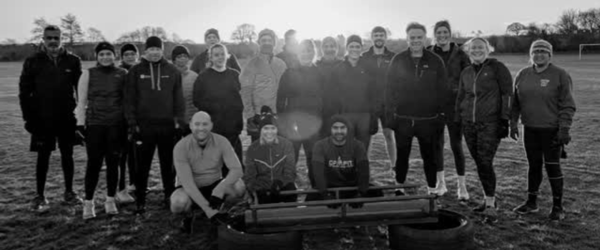 Black and white photo of a group of people outdoors, some standing and some kneeling, on a grassy field with trees in the background.