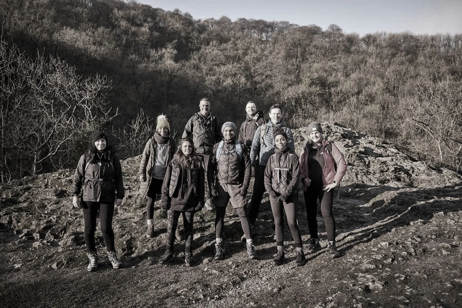 Group of nine hikers standing on rocky terrain outdoors with trees and hills in the background.