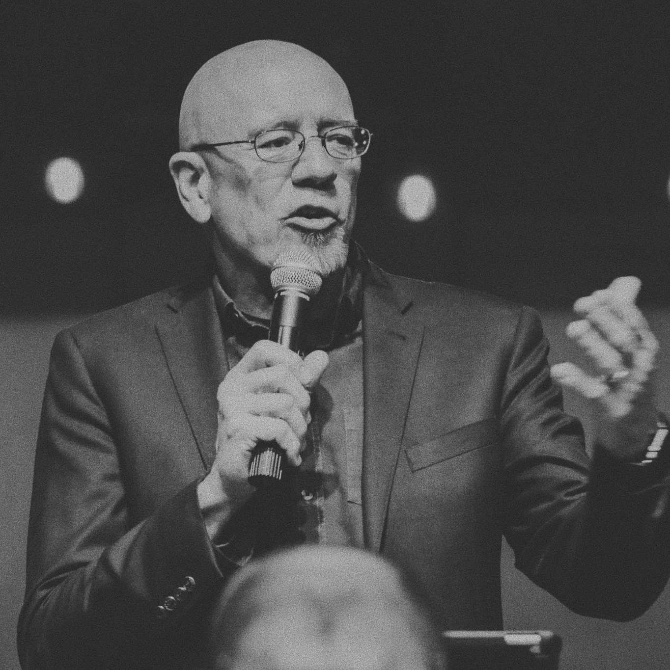 A black and white photograph of a bald man with glasses speaking into a microphone, gesturing with his hand, wearing a suit jacket.