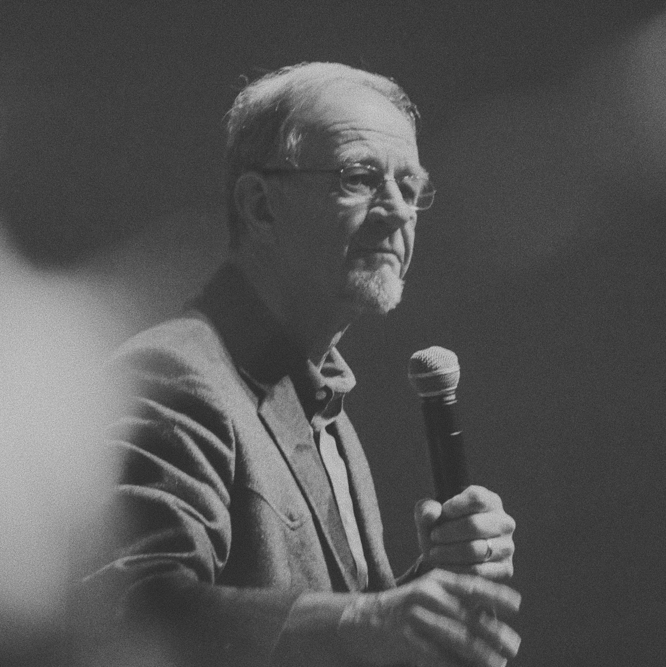 A black and white photo of an older man with glasses, holding a microphone, looking serious.